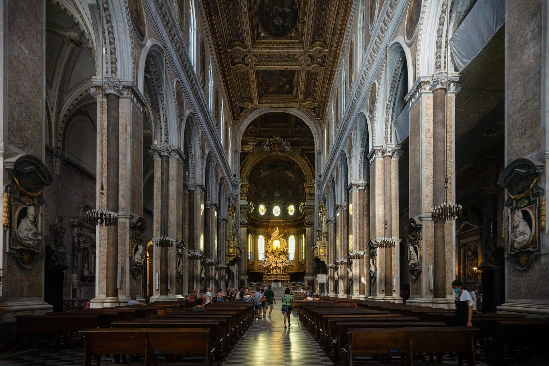 Nave central da Catedral de Nápoles com majestosas colunas de mármore, arcos ornamentados e visitantes admirando o altar iluminado sob uma suave luz diurna.