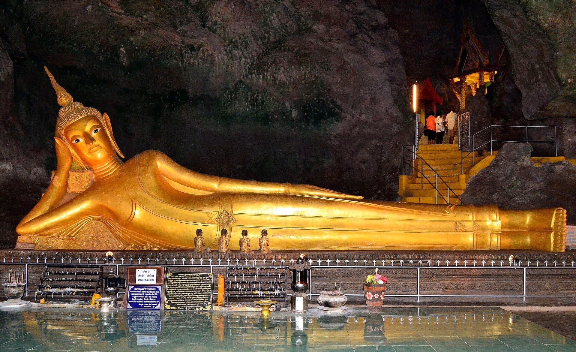 Large golden reclining Buddha statue inside Wat Suwan Kuha cave temple, with rock walls and offerings visible in soft interior lighting.