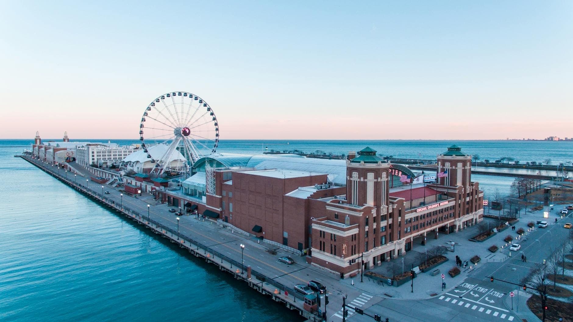 Aerial view of Navy Pier with the Centennial Wheel prominently featuring against Lake Michigan and Chicago’s skyline, captured during daylight with clear blue water and sky.