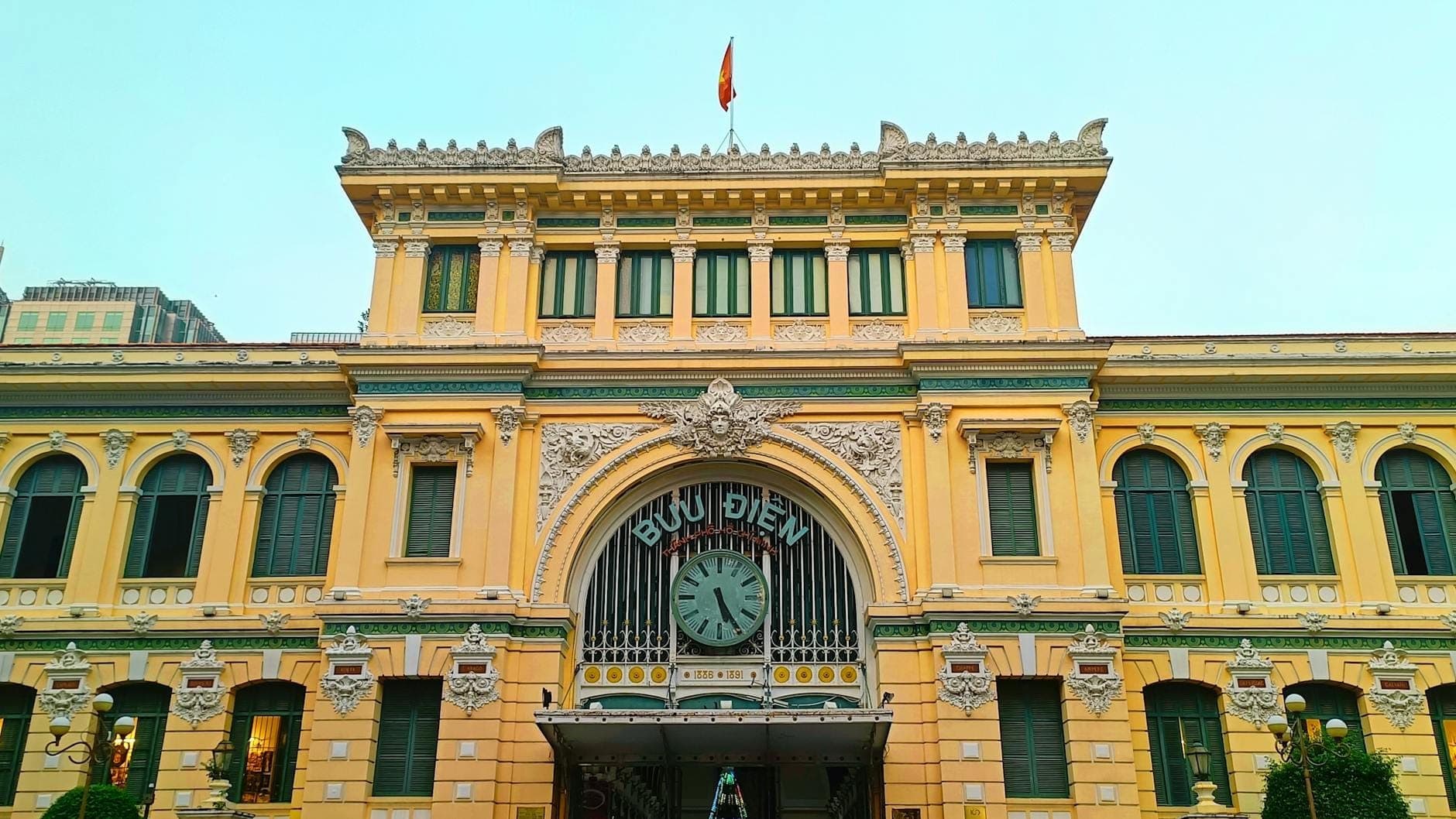 Vista frontal dos Correios Centrais de Saigon, um ornamentado edifício colonial francês amarelo com relógio central e bandeira, sob um céu limpo.