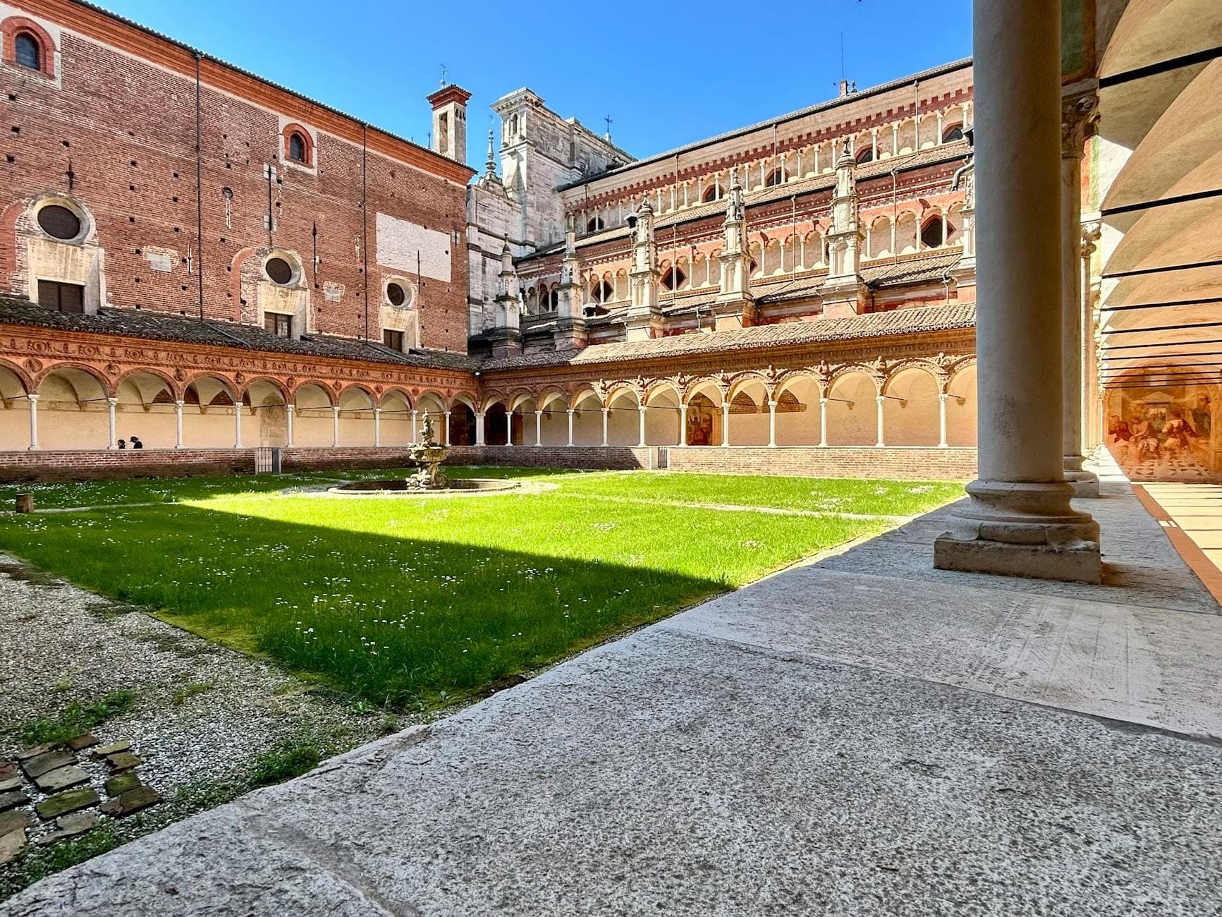 Vista do claustro sereno da Certosa di San Martino com corredores arqueados, uma fonte central e paredes históricas de tijolo sob um céu azul limpo.