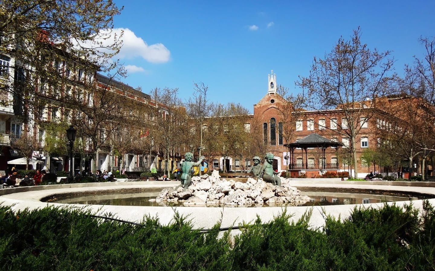 Plaza de Olavide ที่เต็มไปด้วยแสงแดดใน Chamberí กรุงมาดริด โดดเด่นด้วยน้ำพุกลางลานที่มีรูปปั้นสำริด ล้อมรอบด้วยต้นไม้ ม้านั่ง และอาคารอพาร์ตเมนต์สไตล์คลาสสิกในศตวรรษที่ 19