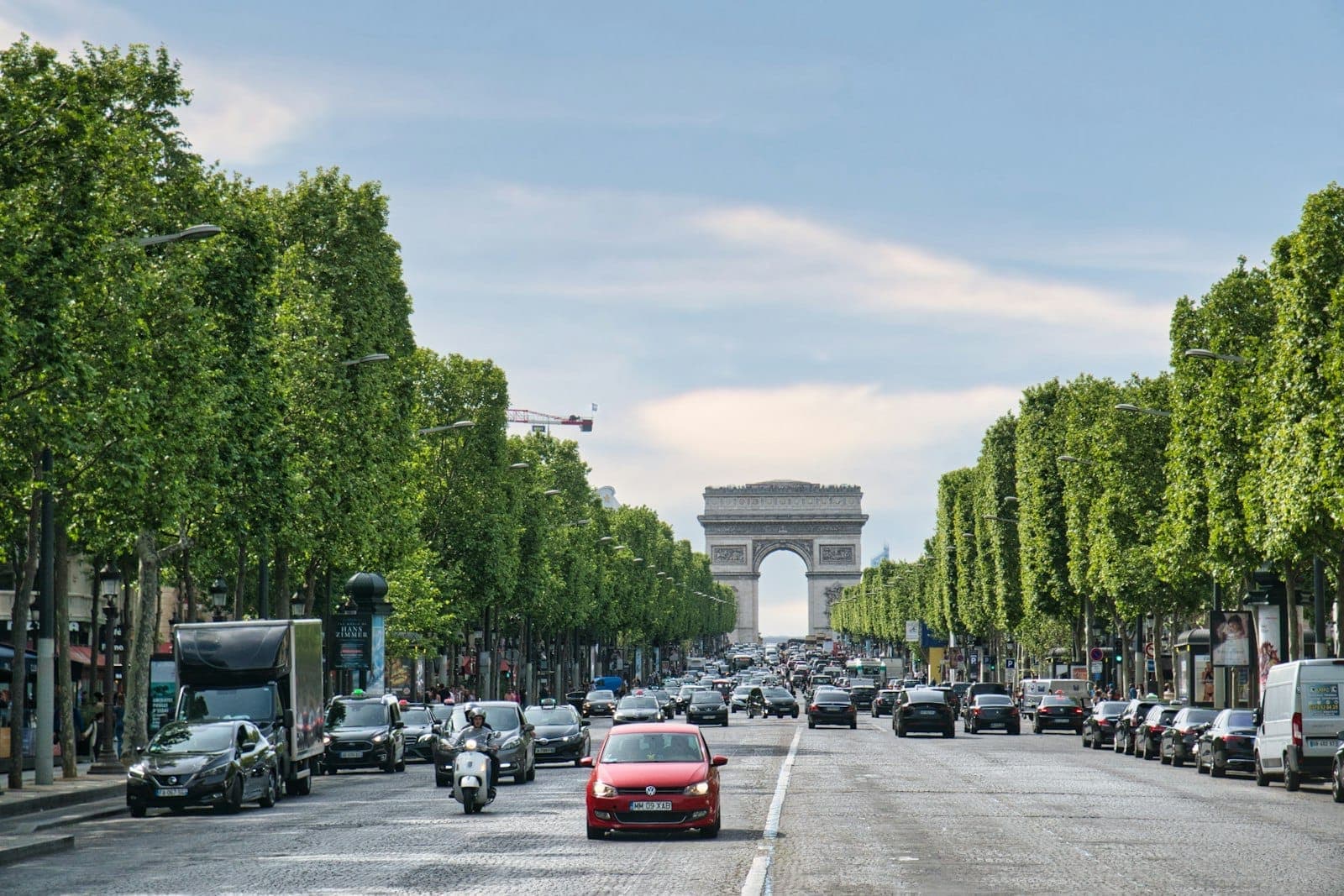 Wide view of the Champs-Élysées leading toward the Arc de Triomphe, lined with green trees and busy with cars under a bright sky.