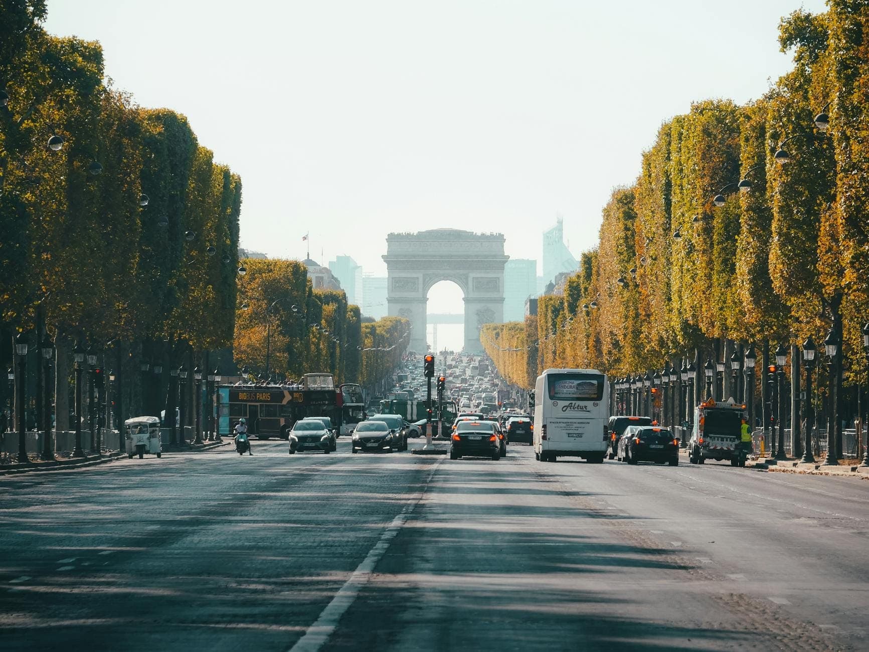 A wide view down the Champs-Élysées lined with trees and traffic, leading towards the Arc de Triomphe on a clear day.