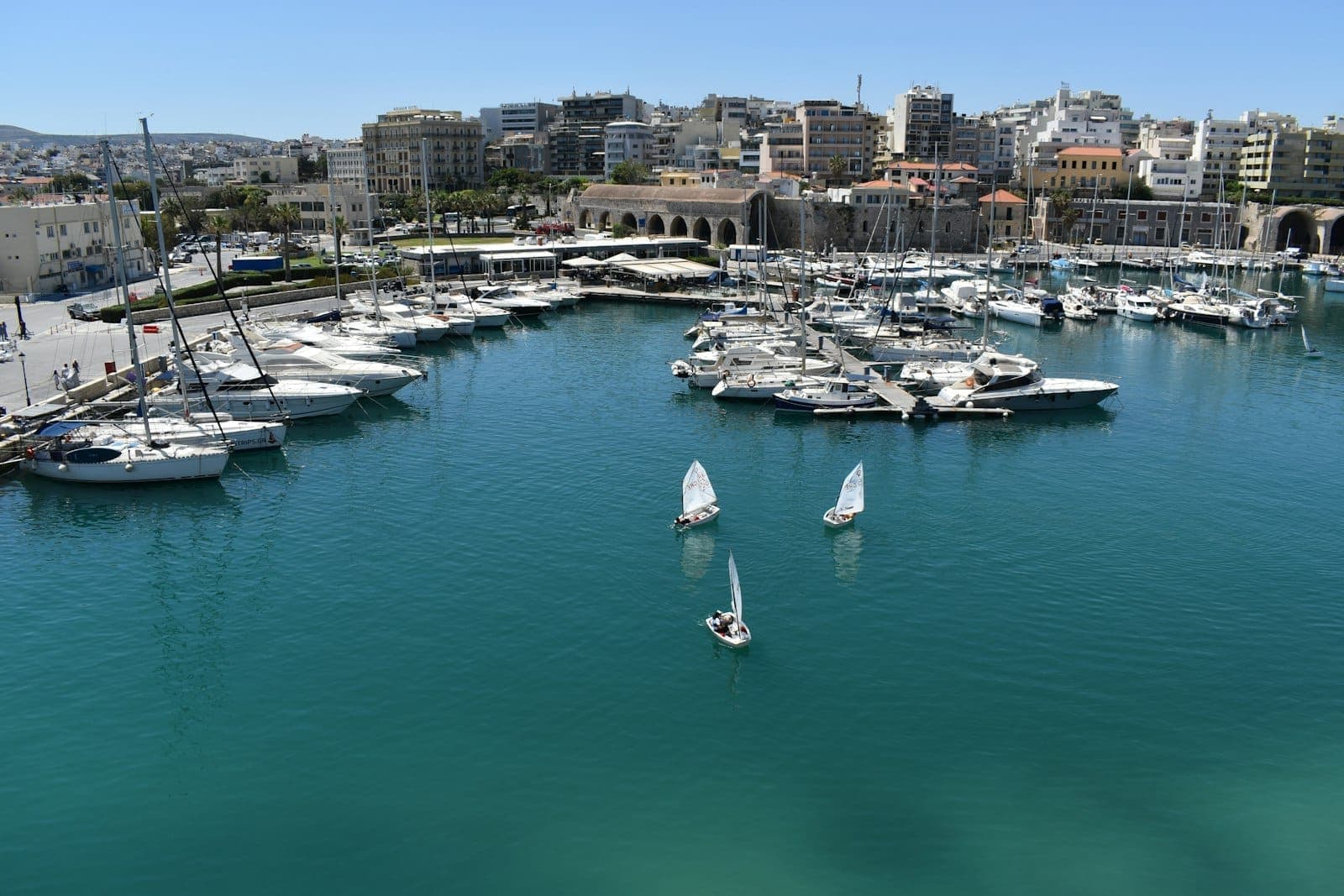 Blick auf den Hafen von Heraklion mit modernen Stadtgebäuden, Booten und Yachten. Städtische Uferszene unter einem klaren blauen Himmel.