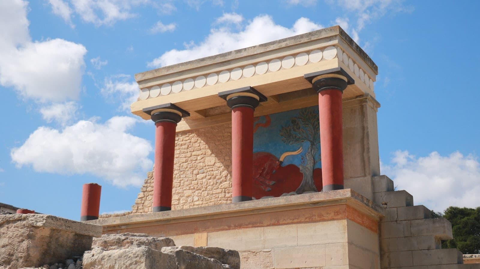 Gut erhaltene Ruinen des Palastes von Knossos in Heraklion, mit markanten roten Säulen und einer Rekonstruktion eines farbenfrohen minoischen Freskos.