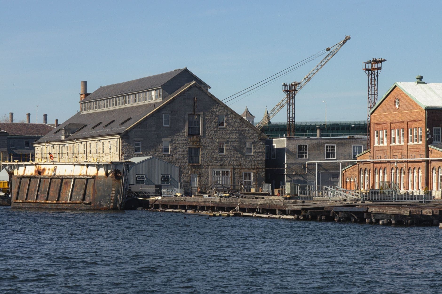 Bâtiments historiques en pierre et en brique sur le front de mer du Charlestown Navy Yard, avec une grue et une barge à quai, témoins de l'héritage maritime et industriel du site.