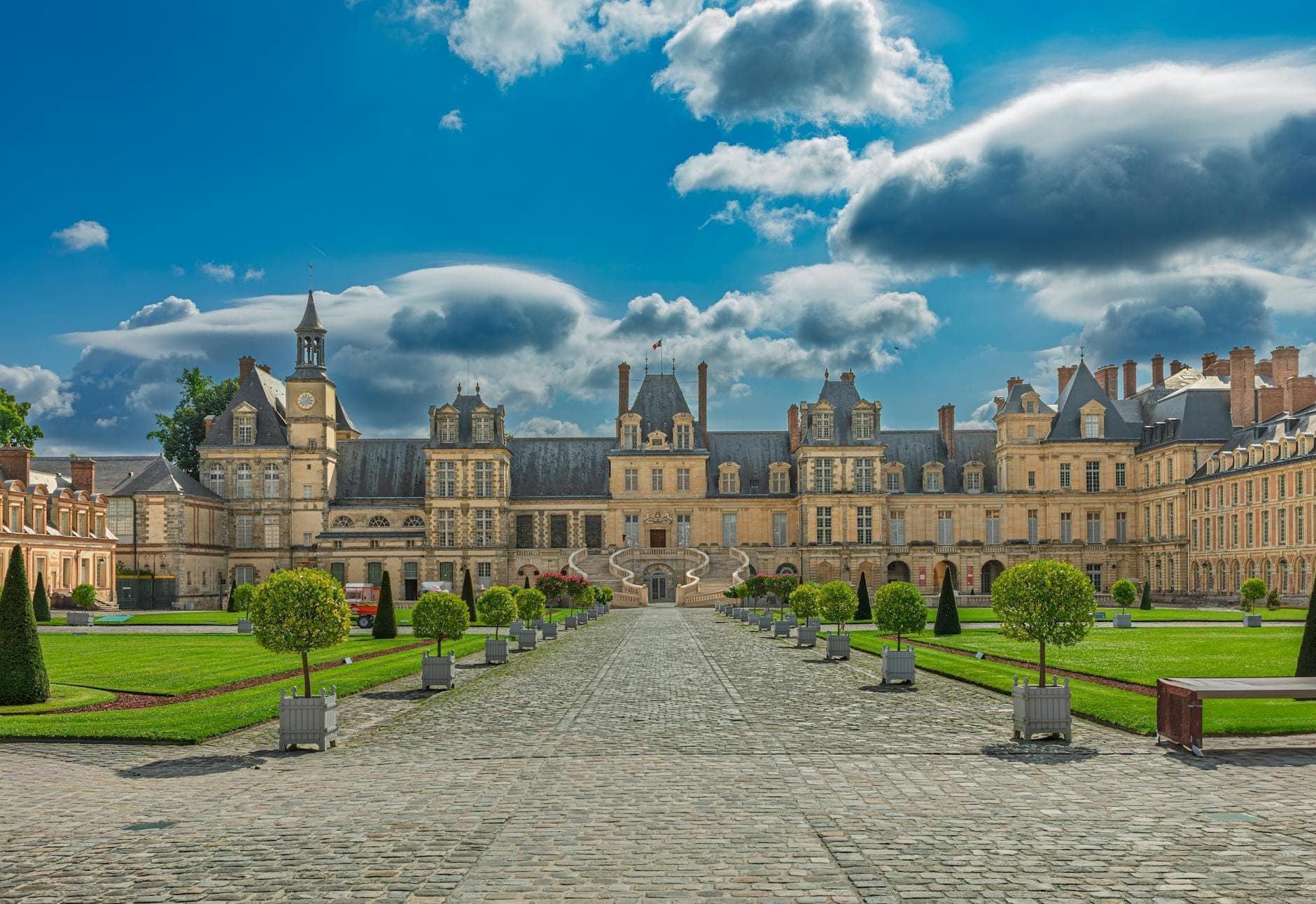 Wide-angle view of Château de Fontainebleau’s grand façade, formal gardens, and blue sky with dramatic clouds, capturing the palace’s grandeur and inviting entrance approach.
