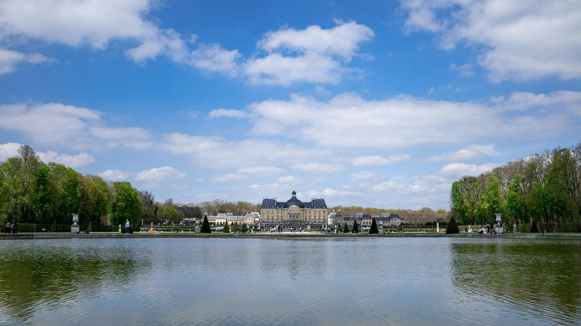 The Château de Vaux-le-Vicomte viewed across a large reflective pond, surrounded by formal gardens, all under a bright blue sky with scattered clouds.