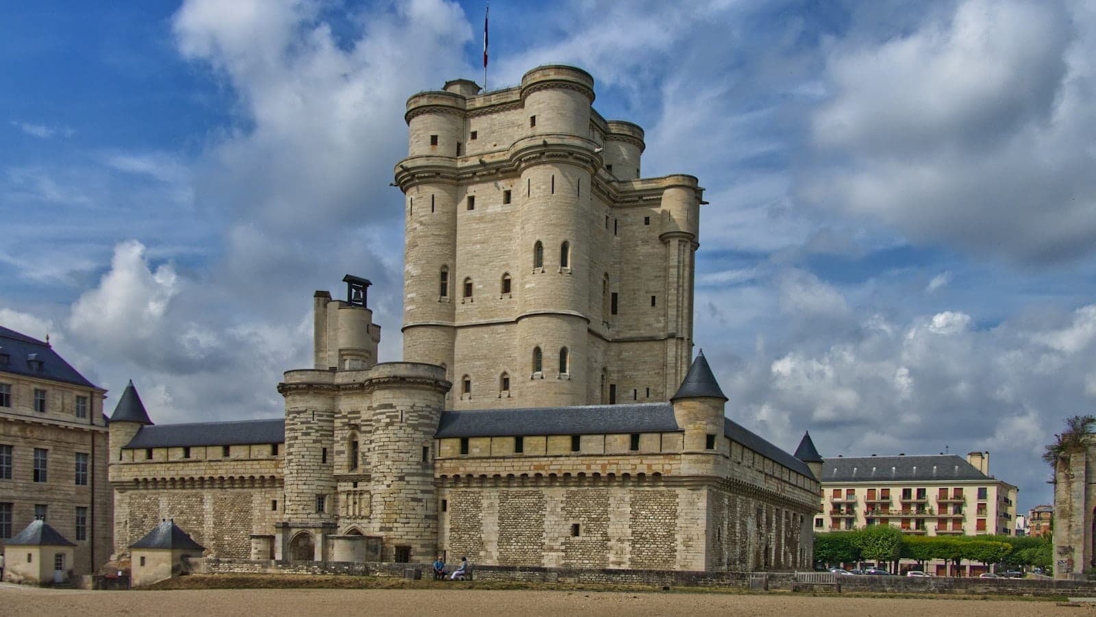 Wide view of Château de Vincennes with its imposing medieval keep, turrets, and fortress walls under a dramatic blue sky with scattered clouds.