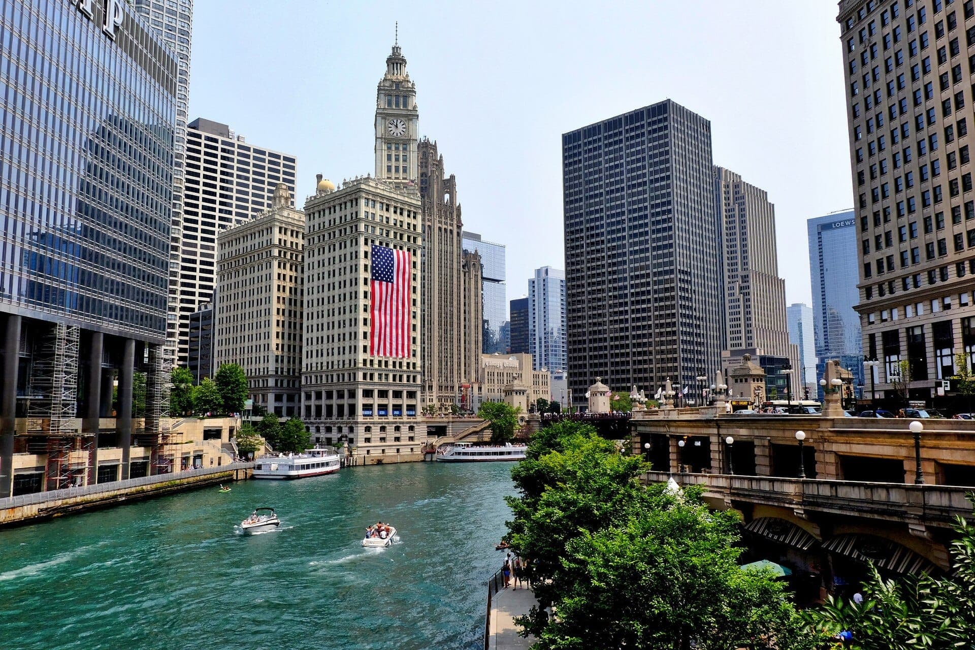 The Chicago River flows past downtown skyscrapers, including the Wrigley Building with a large American flag displayed for July 4th celebrations.