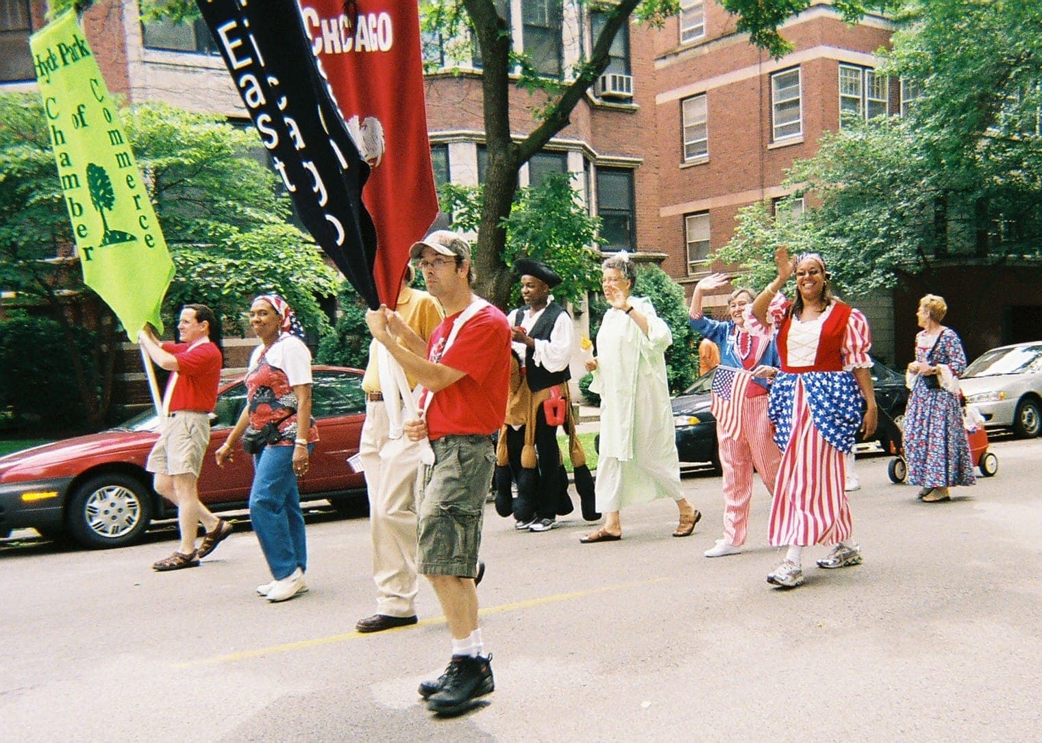 People in festive red, white, and blue costumes walk in a neighborhood parade with banners, brick buildings, and leafy trees lining the street.