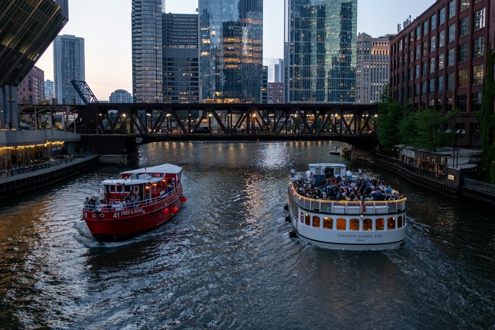 Two tour boats filled with people cruise the Chicago River at dusk with illuminated skyscrapers and a bridge in view.