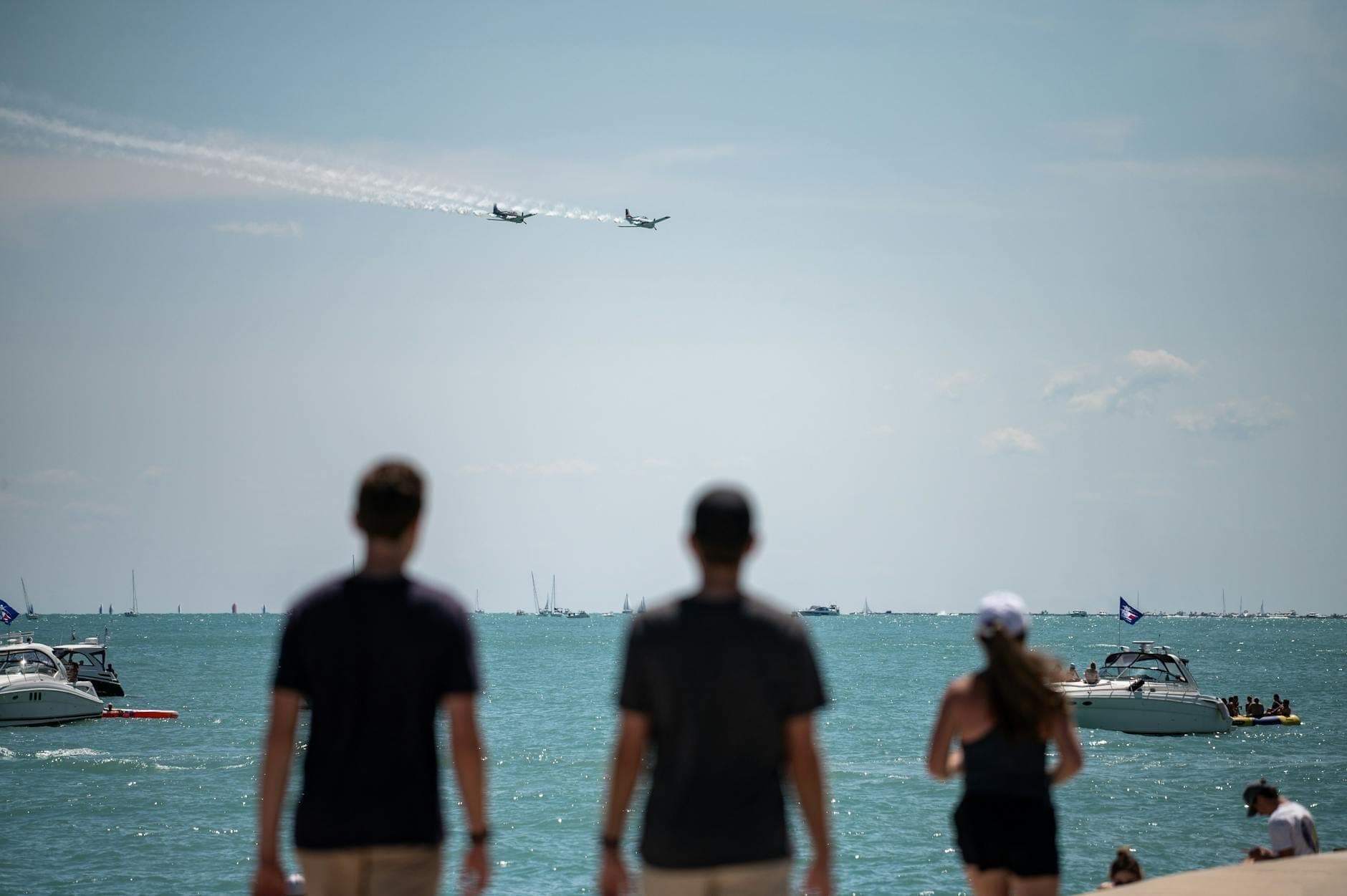 Personas en el frente del lago de Chicago observan cómo los aviones realizan maniobras aéreas sobre el lago Míchigan, con embarcaciones en el agua en un día soleado.
