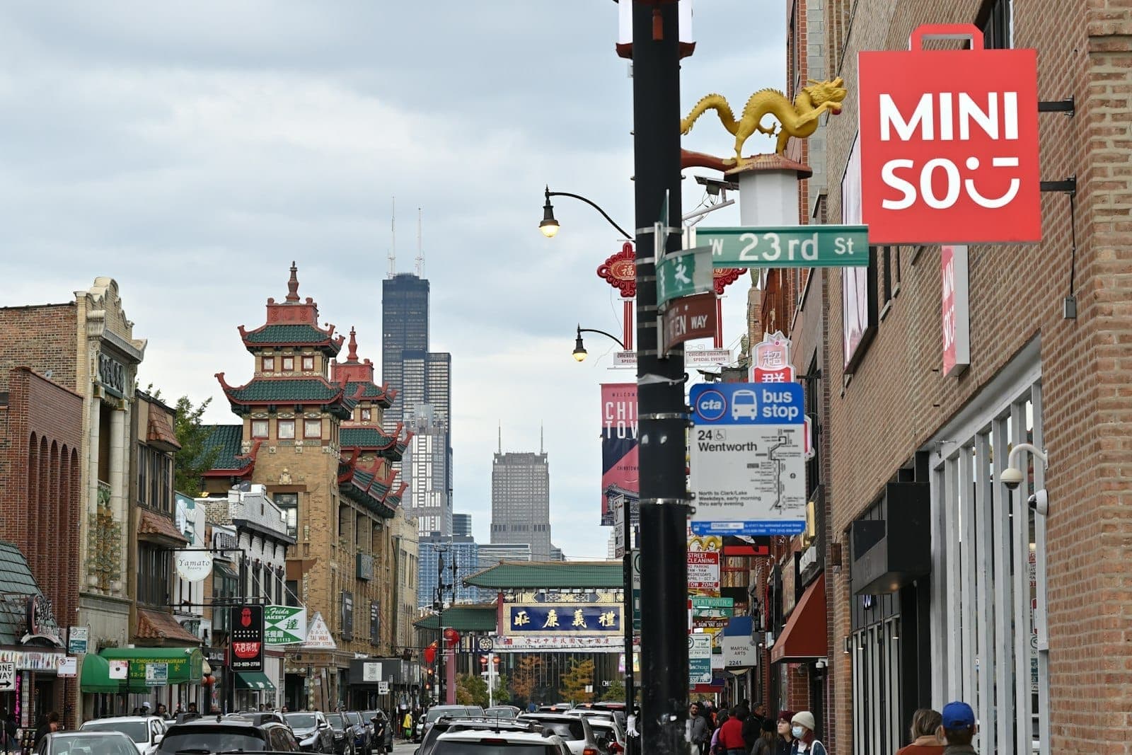 Street view of Chicago’s Chinatown with Chinese-style buildings, colorful signage, and the downtown skyline in the background on a cloudy day.
