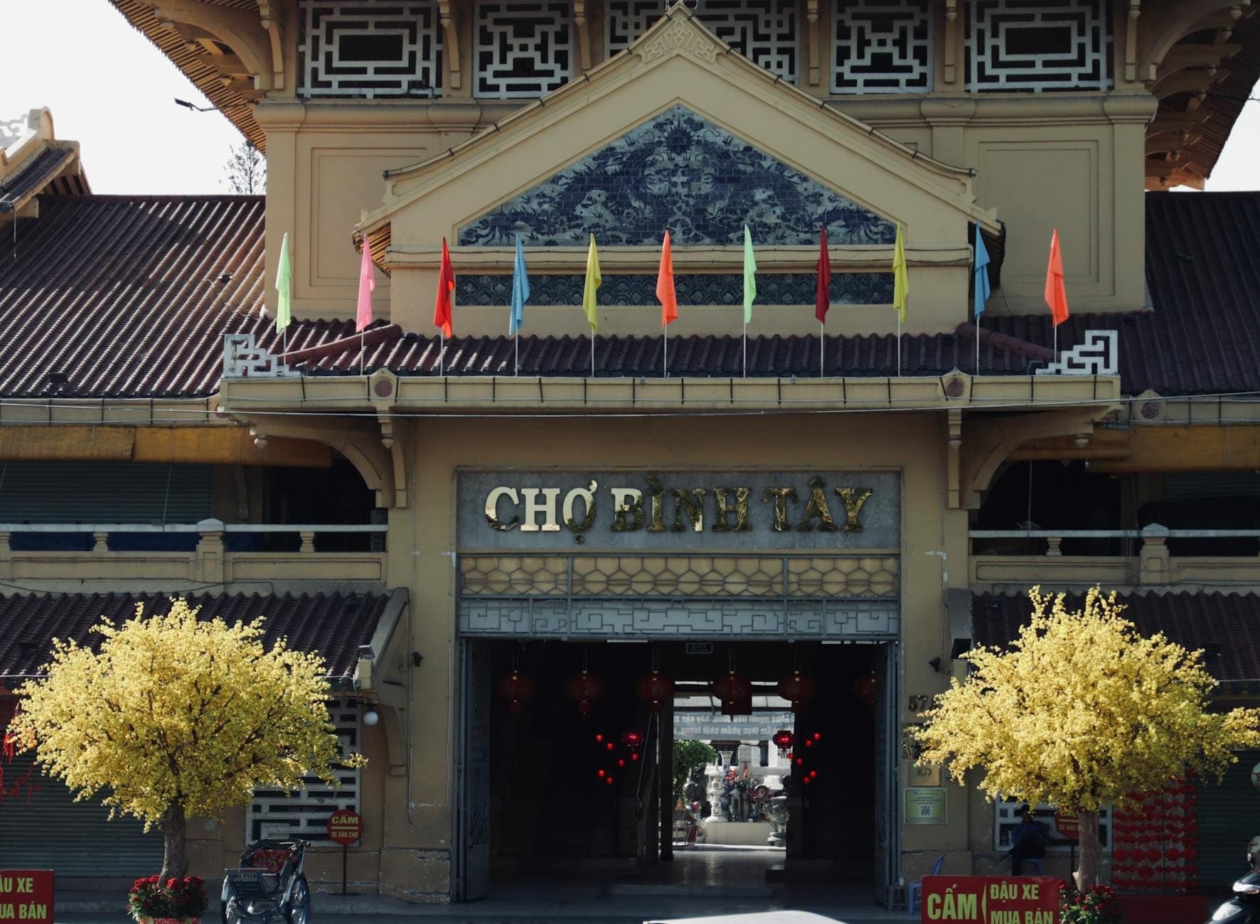 Фасад исторического рынка Чợ Бình Тây в Чợ Лớн, украшенный разноцветными флагами и жёлтыми цветущими деревьями.