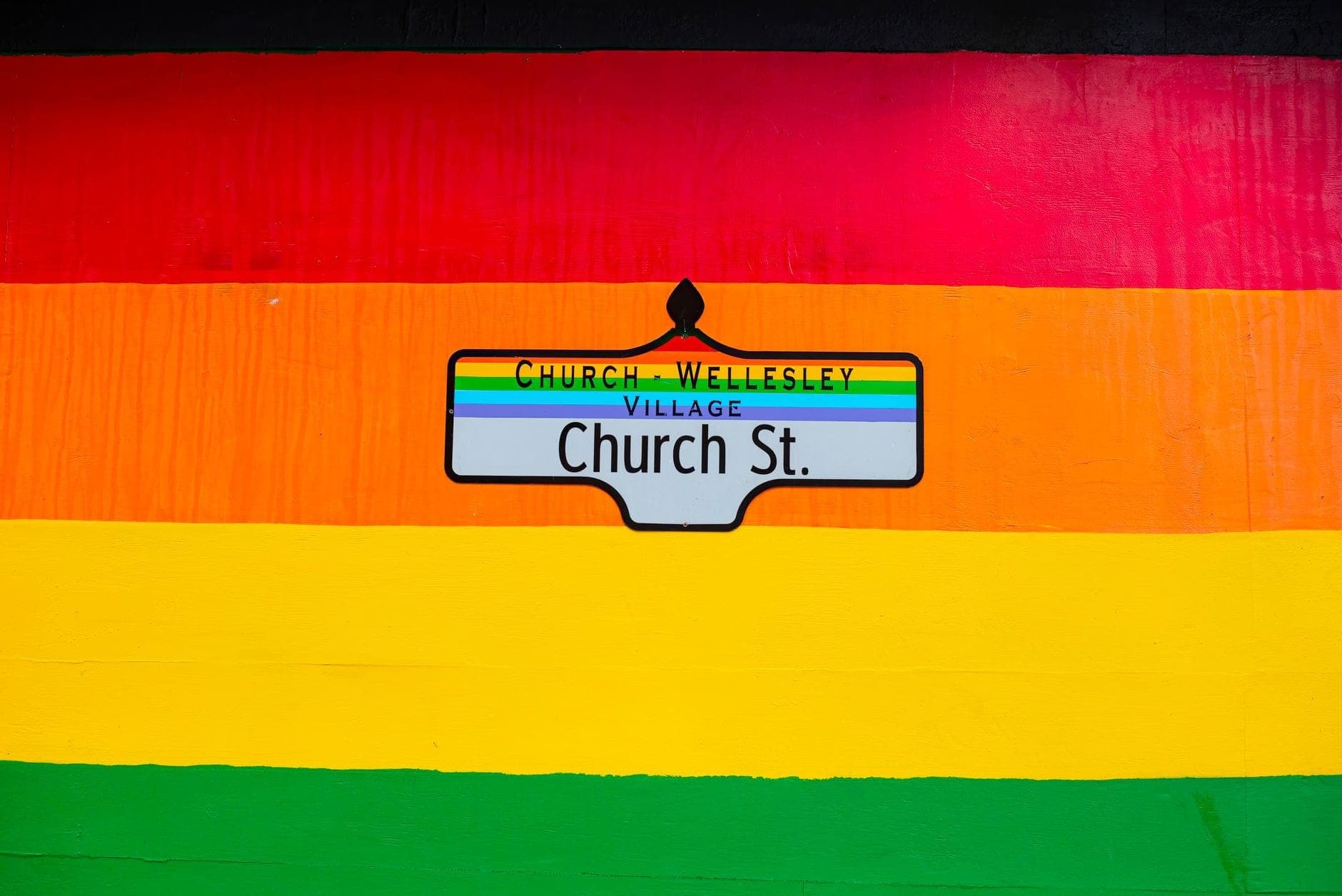 Un vivace murale arcobaleno con il cartello stradale del Church-Wellesley Village al centro, simbolo del quartiere LGBTQ+ di Toronto e del suo spirito inclusivo e colorato.