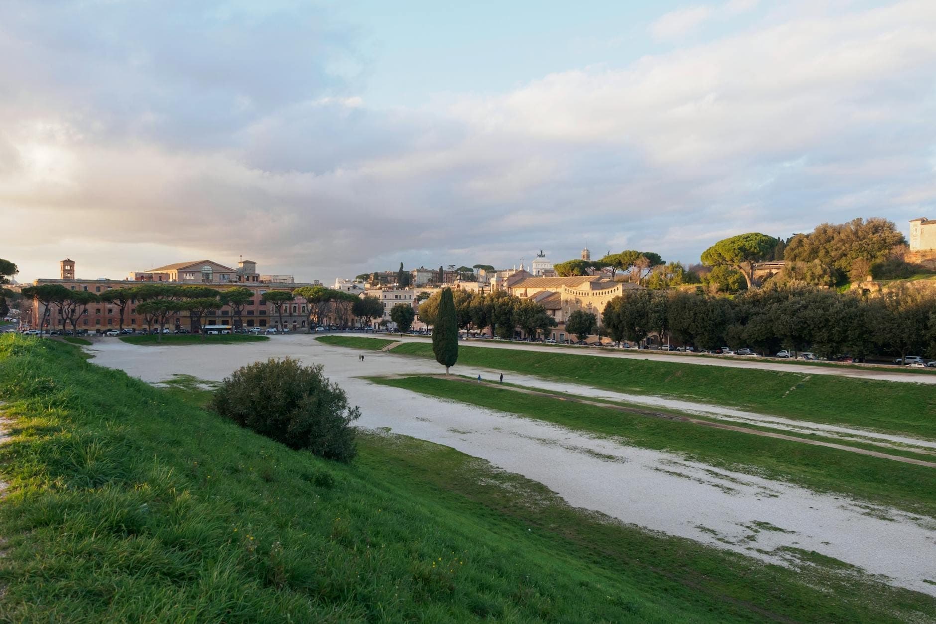 Vista panoramica del Circo Massimo a Roma, con il profilo dell'antico stadio dei carri, i pendii erbosi, il cielo aperto e i palazzi della città sullo sfondo.