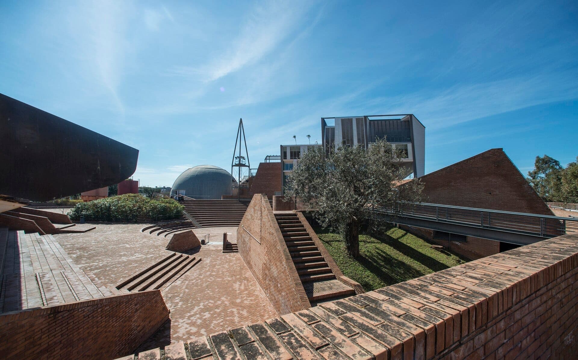 Fachada moderna da Città della Scienza em Nápoles com caminhos de tijolos, degraus de anfiteatro, área verde e a icônica cúpula do planetário em um dia ensolarado.