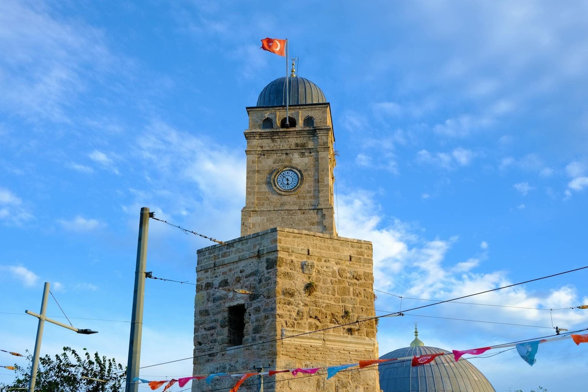 Osmanischer Steinuhrturm Saat Kulesi in Antalya mit türkischer Flagge auf der Spitze, hellem Himmel im Hintergrund und festlichen Wimpeln im Vordergrund.
