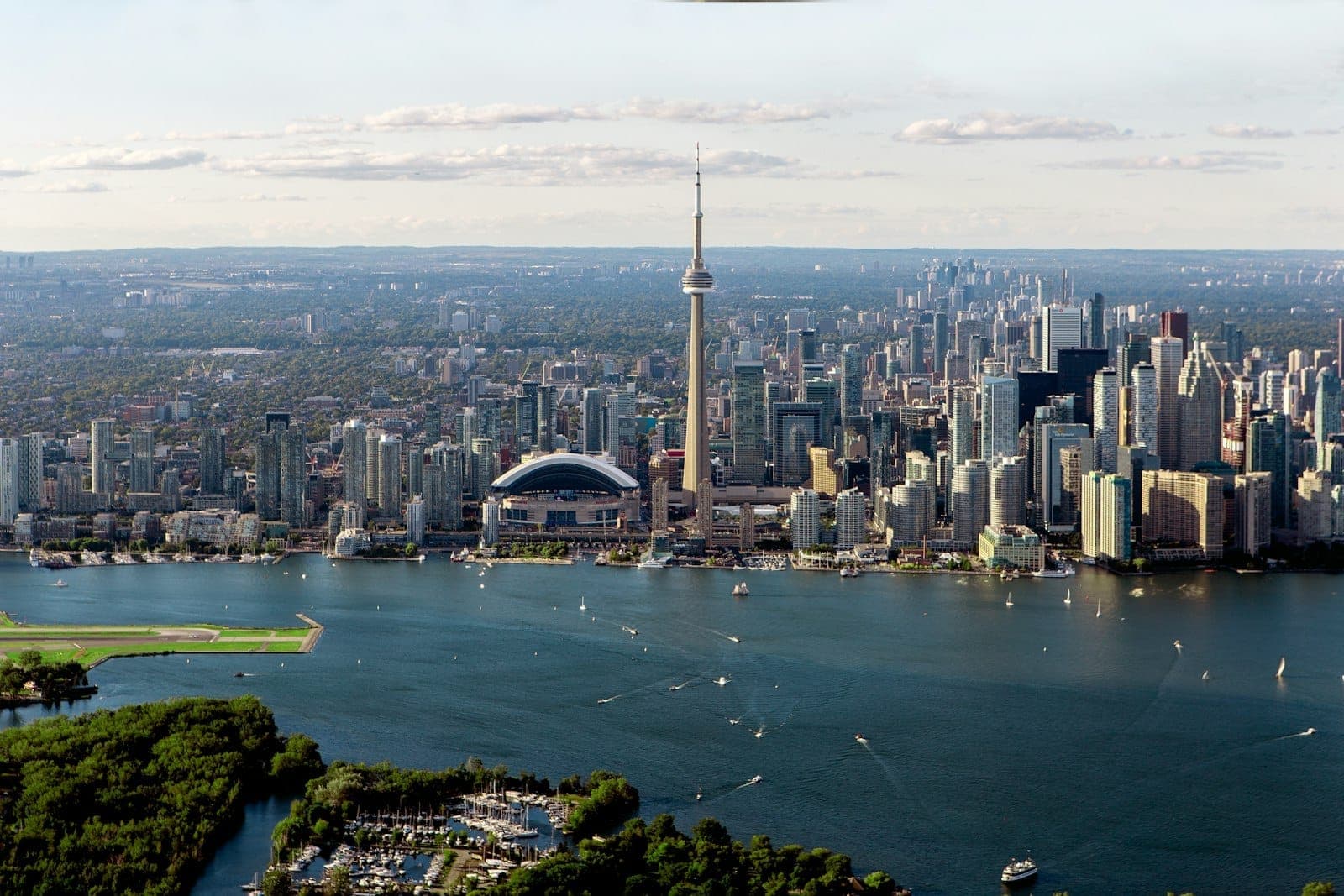 Vista aerea panoramica del centro di Toronto con la CN Tower che svetta sullo skyline della città, il Lago Ontario e alcune imbarcazioni in primo piano in una giornata serena.