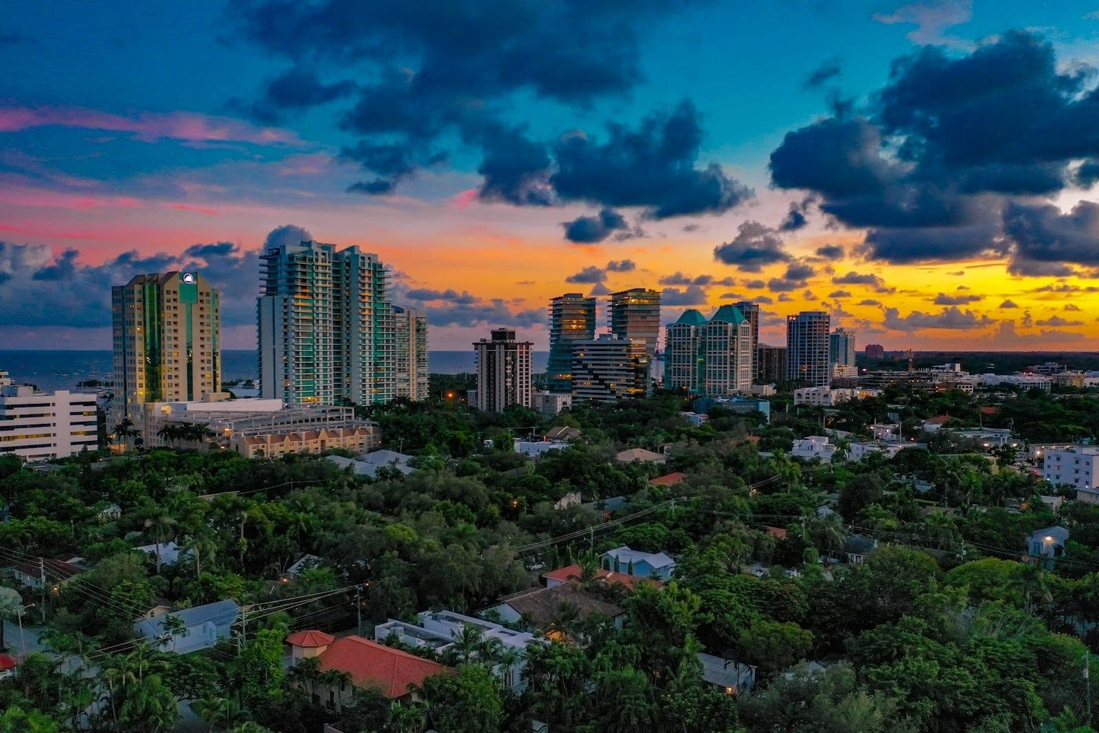 Aerial view of Coconut Grove at sunset, showing lush greenery, banyan trees, mid-rise buildings, and Biscayne Bay in the background under a vibrant sky.