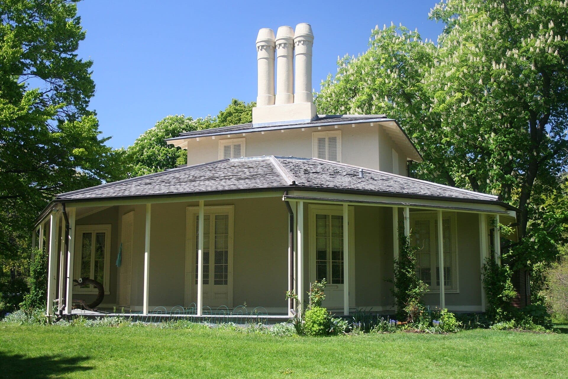 Museo storico Colborne Lodge, un edificio in stucco beige con camini unici, circondato da rigogliosi alberi verdi sotto un cielo azzurro brillante.