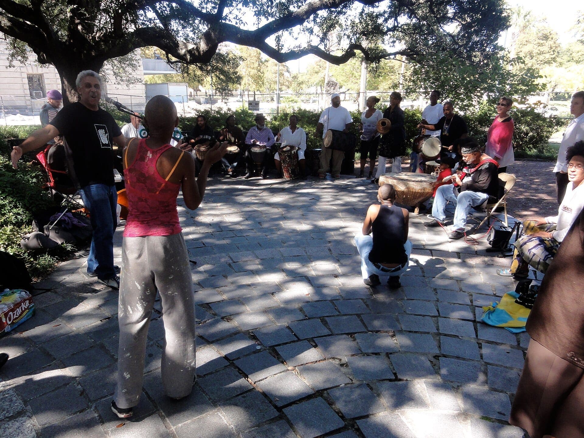 A group of people gathers under a large oak tree at Congo Square, drumming, dancing, and playing music on a sunny day.
