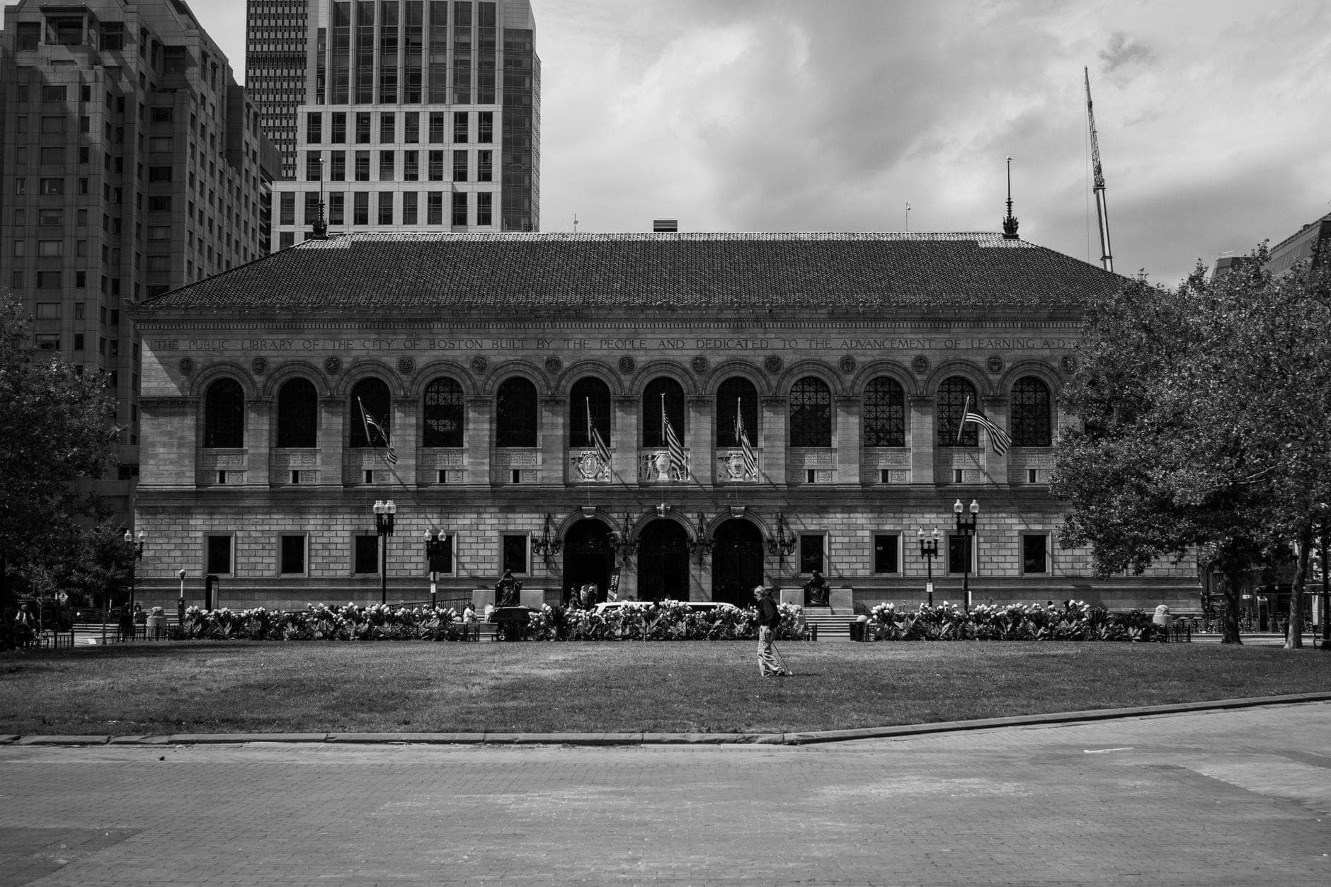Vue frontale de la historic Boston Public Library à Copley Square, entourée de gratte-ciel, avec des passants et de la végétation au premier plan.