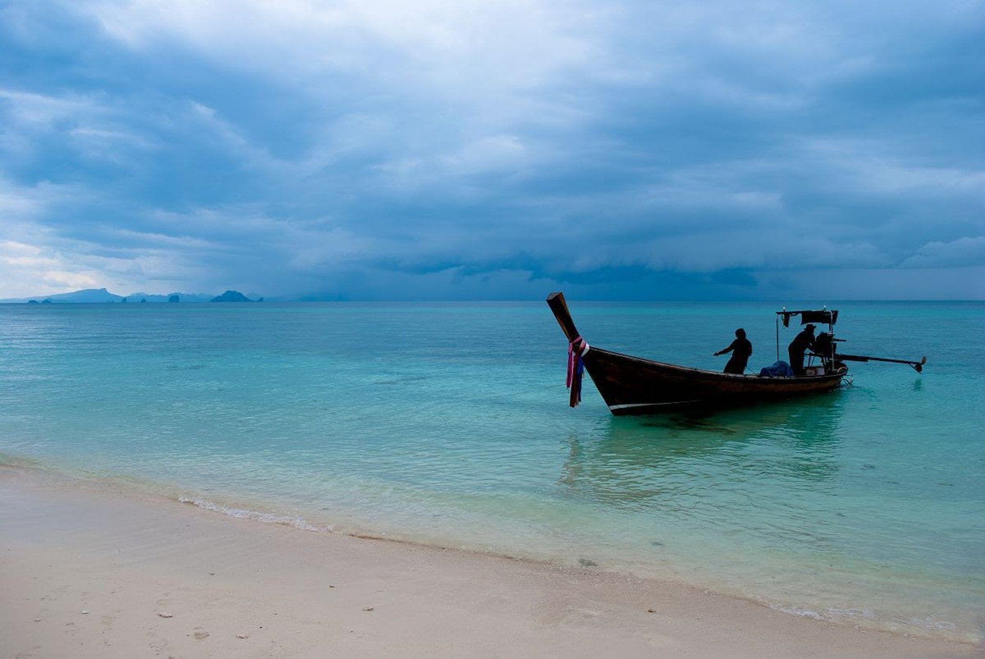 Traditional long-tail boat anchored in clear turquoise waters off Coral Island, Phuket, with sandy beach and dramatic blue sky overhead.