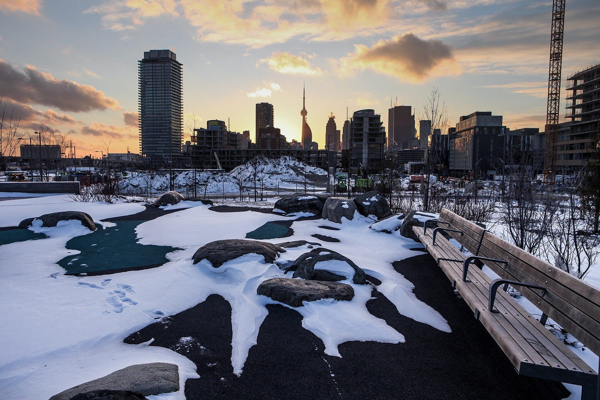 Corktown Common in inverno, coperto da chiazze di neve, grandi rocce, panchine di legno e lo skyline di Toronto silhouettato al tramonto.
