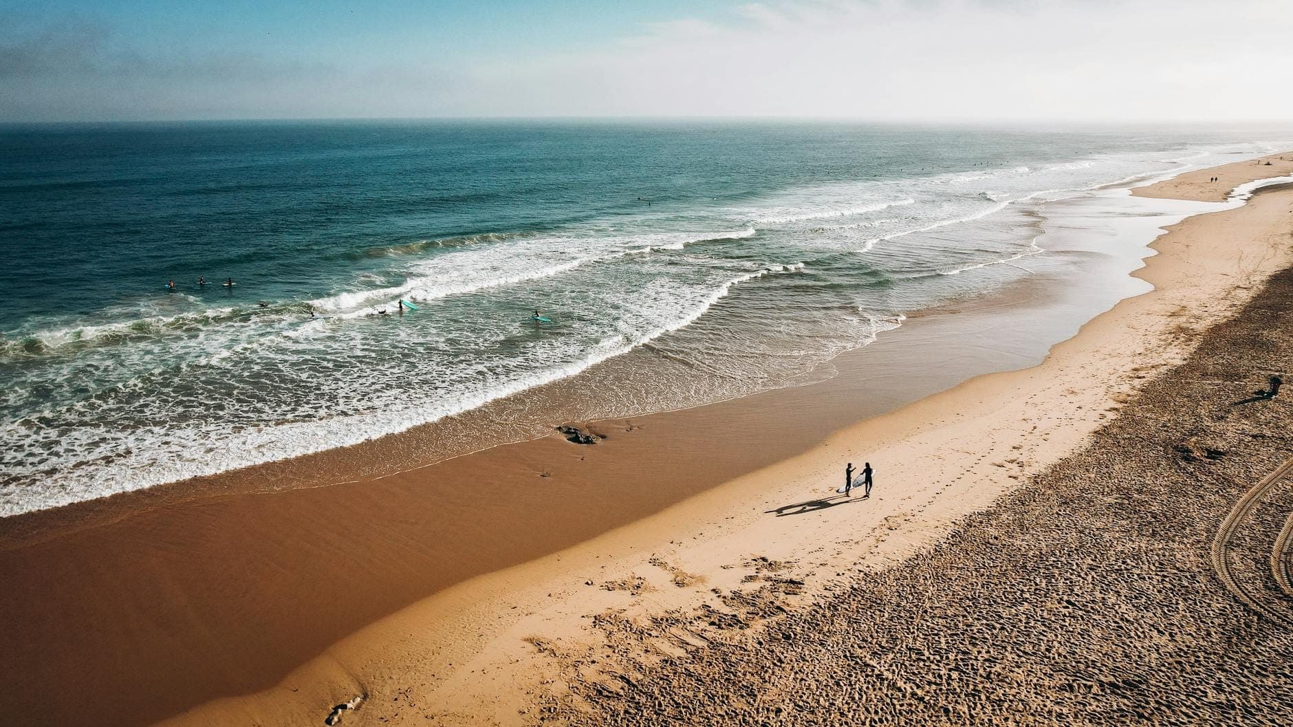 Vista aérea de uma longa faixa de areia com ondas turquesa e algumas pessoas caminhando à beira-mar sob um céu luminoso.