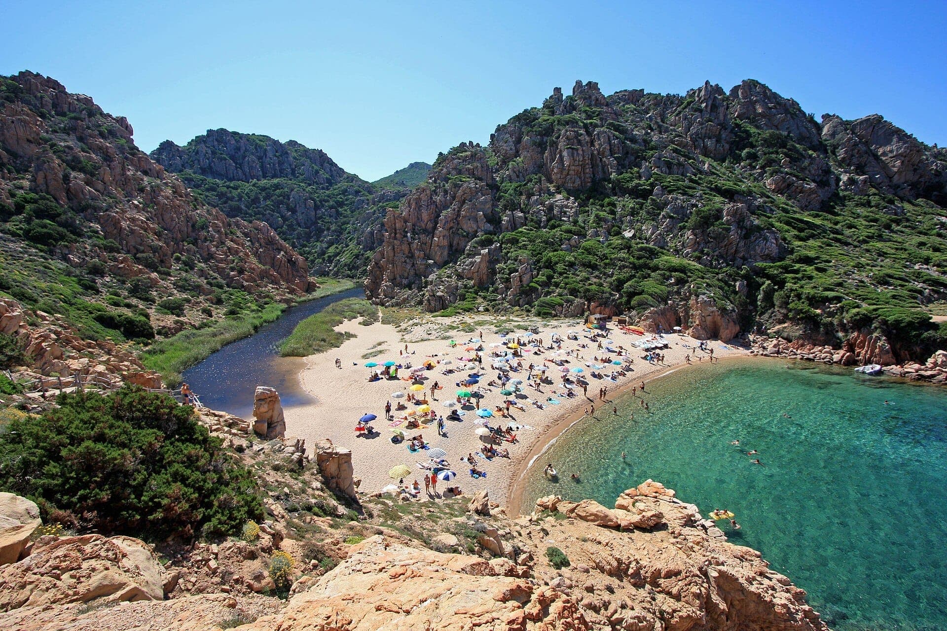 Sandy beach at Costa Paradiso surrounded by rugged red granite cliffs, clear turquoise water, and sunbathers with colorful umbrellas on a bright sunny day.