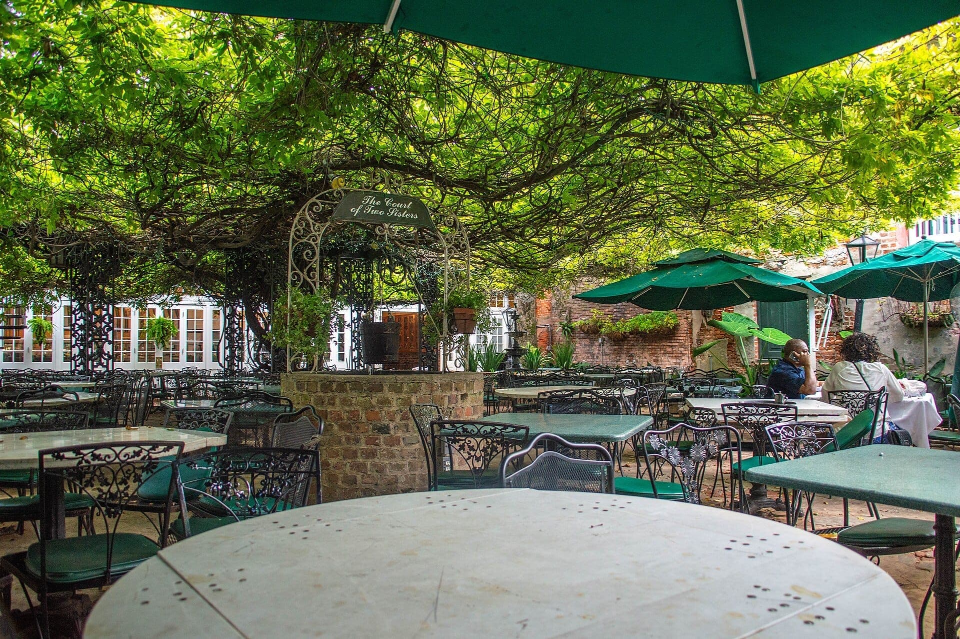 Courtyard dining area at The Court of Two Sisters, featuring wrought-iron tables, green umbrellas, brick well, and lush greenery overhead.