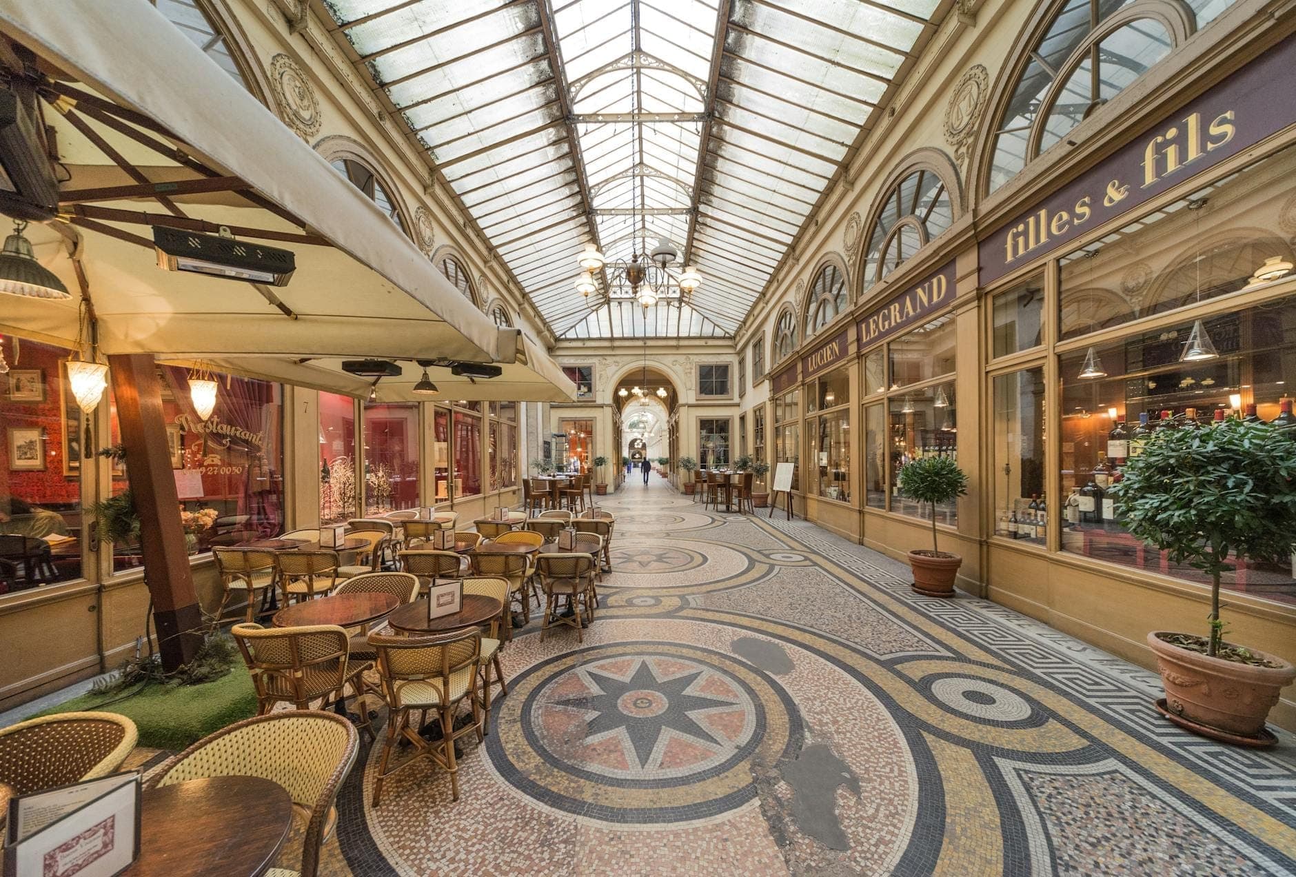 Interior view of a Parisian covered passage with mosaic floors, glass roof, elegant storefronts, and inviting café tables lining the arcade.