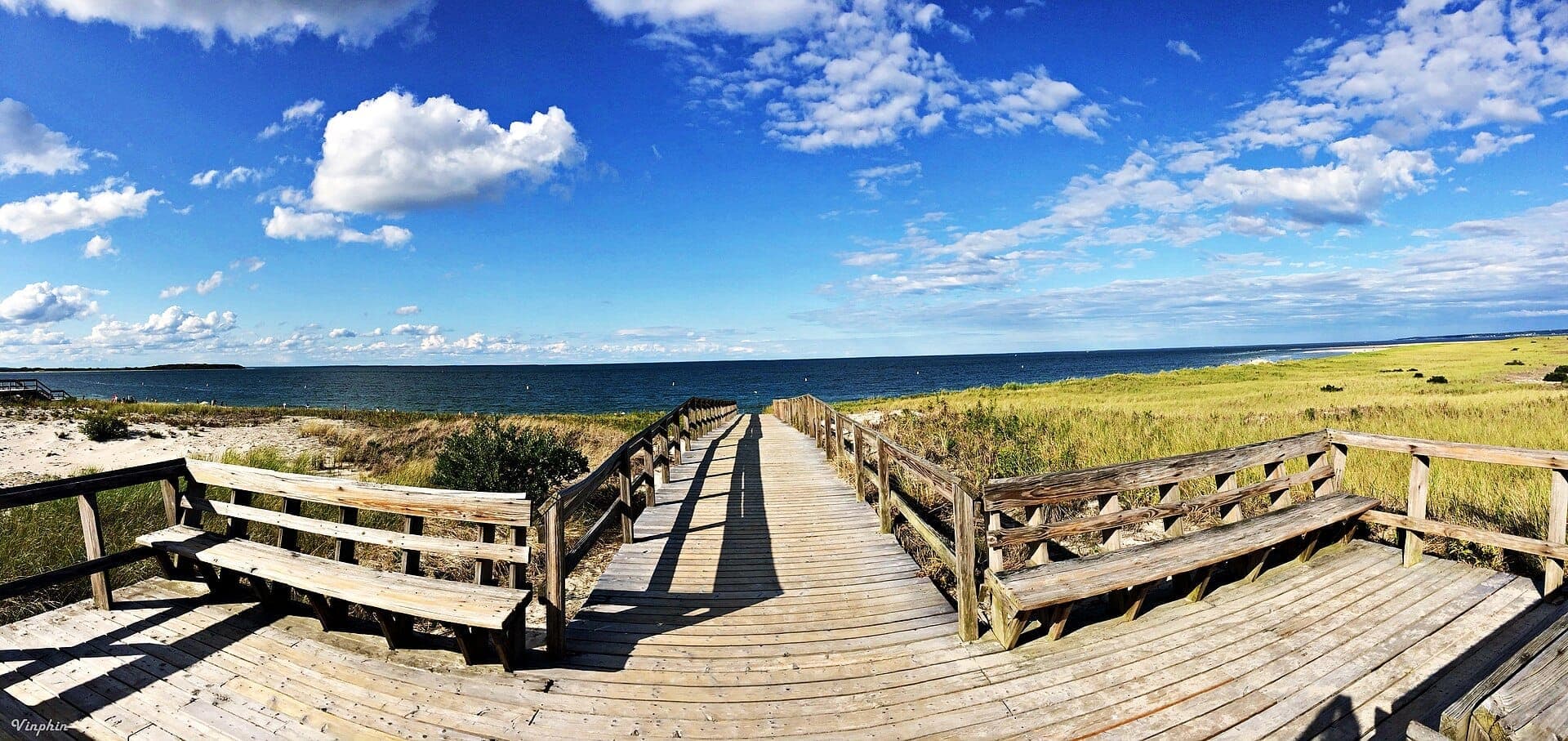 Large promenade en bois et bancs surplombant les dunes de sable et l'eau bleue à Crane Beach, avec un paysage herbeux sous un ciel lumineux.