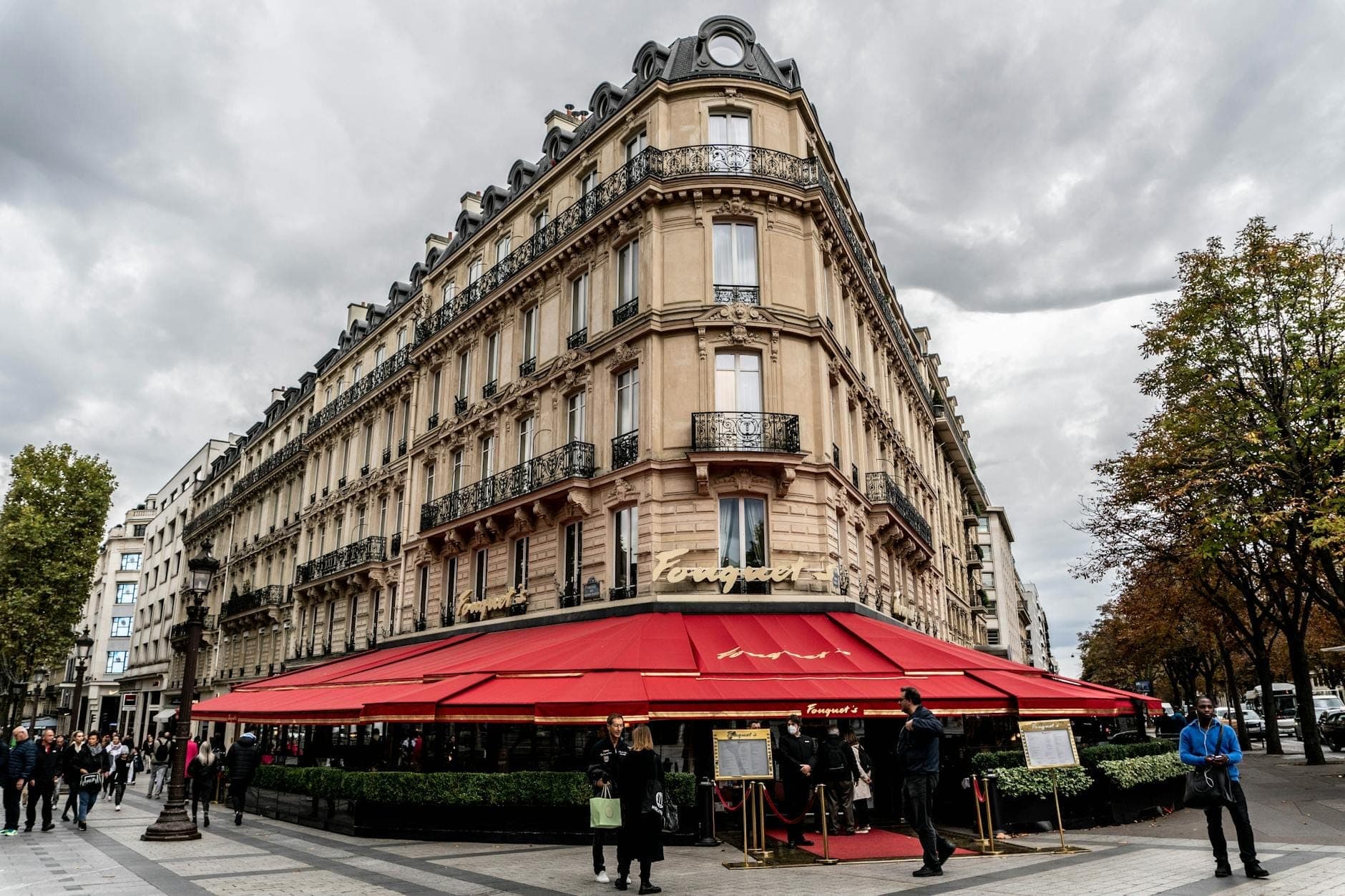 Elegant Parisian building on a busy corner with a red awning, people gathering outside, and trees lining the street under a dramatic cloudy sky.