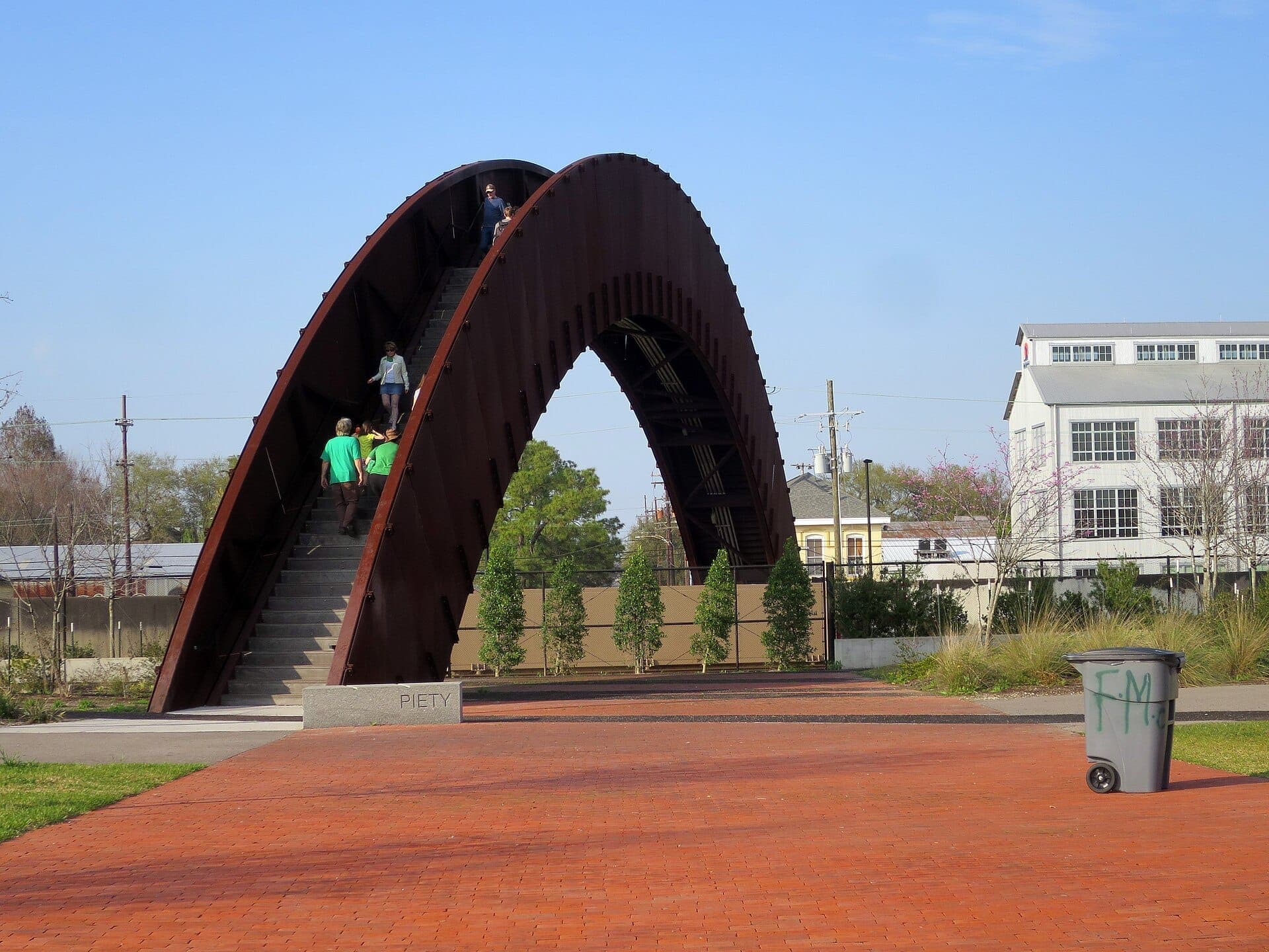 Ponte pedonal no Crescent Park com pessoas subindo as escadas arqueadas, rodeada de vegetação e edifícios do bairro Bywater sob um céu azul limpo.
