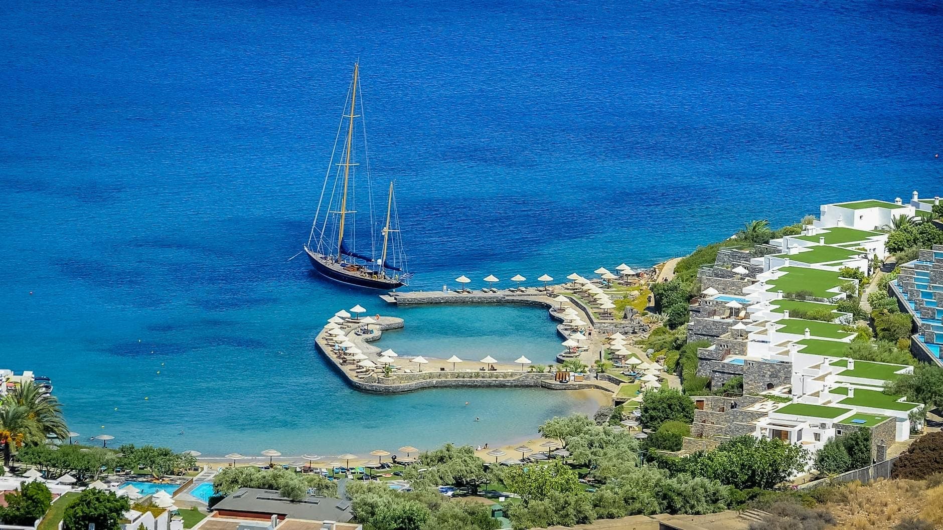 Luftaufnahme eines Strandresorts auf Kreta mit Sonnenliegen, Sonnenschirmen und einem Segelboot in türkisfarbenem Wasser, gehobene Hotelsuiten auf der rechten Seite.