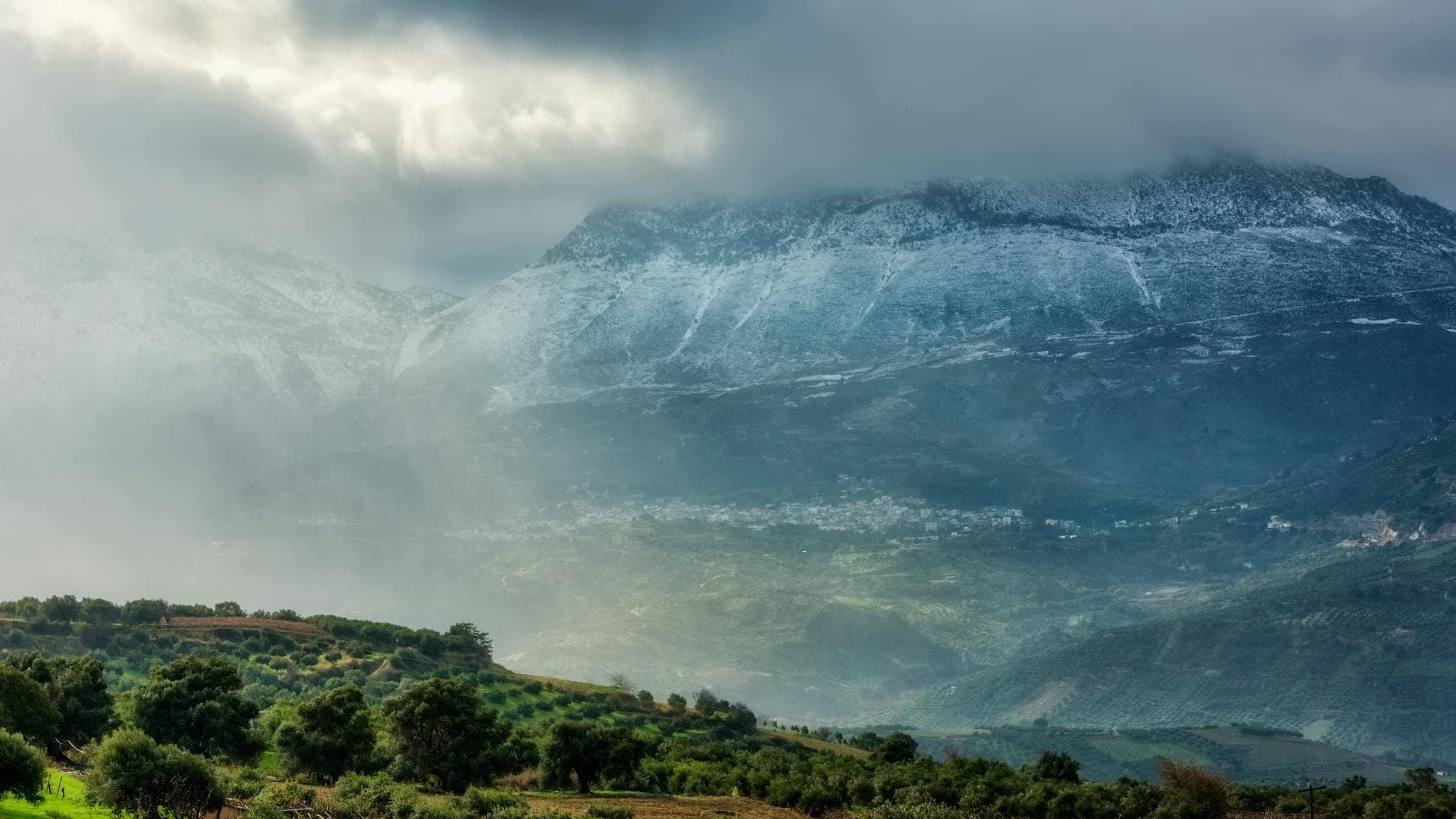 Vista drammatica e nuvolosa delle Montagne Bianche con vette innevate, colline lussureggianti e case di villaggio sparse nell'entroterra occidentale di Creta