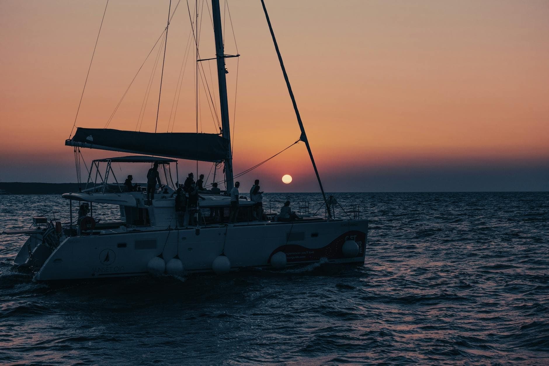 Catamarano con persone che godono di una crociera al tramonto al largo della costa di Creta, con un sole arancione basso sul mare.