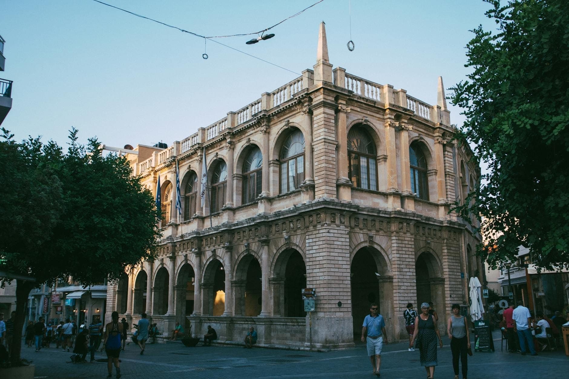 Lo storico edificio della Loggia di Heraklion e la piazza pedonale al crepuscolo, con persone che passeggiano e si radunano nel centro città.