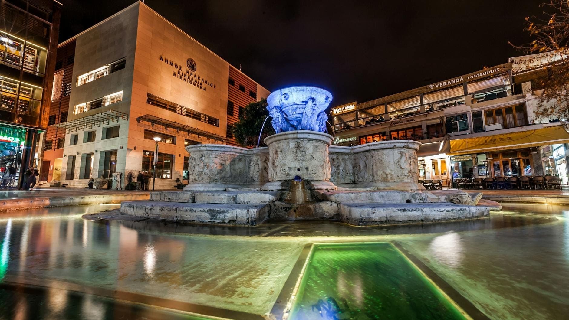 Vista notturna di una famosa piazza di Heraklion con una fontana veneziana illuminata, ristoranti e bar che circondano l'area vivace.