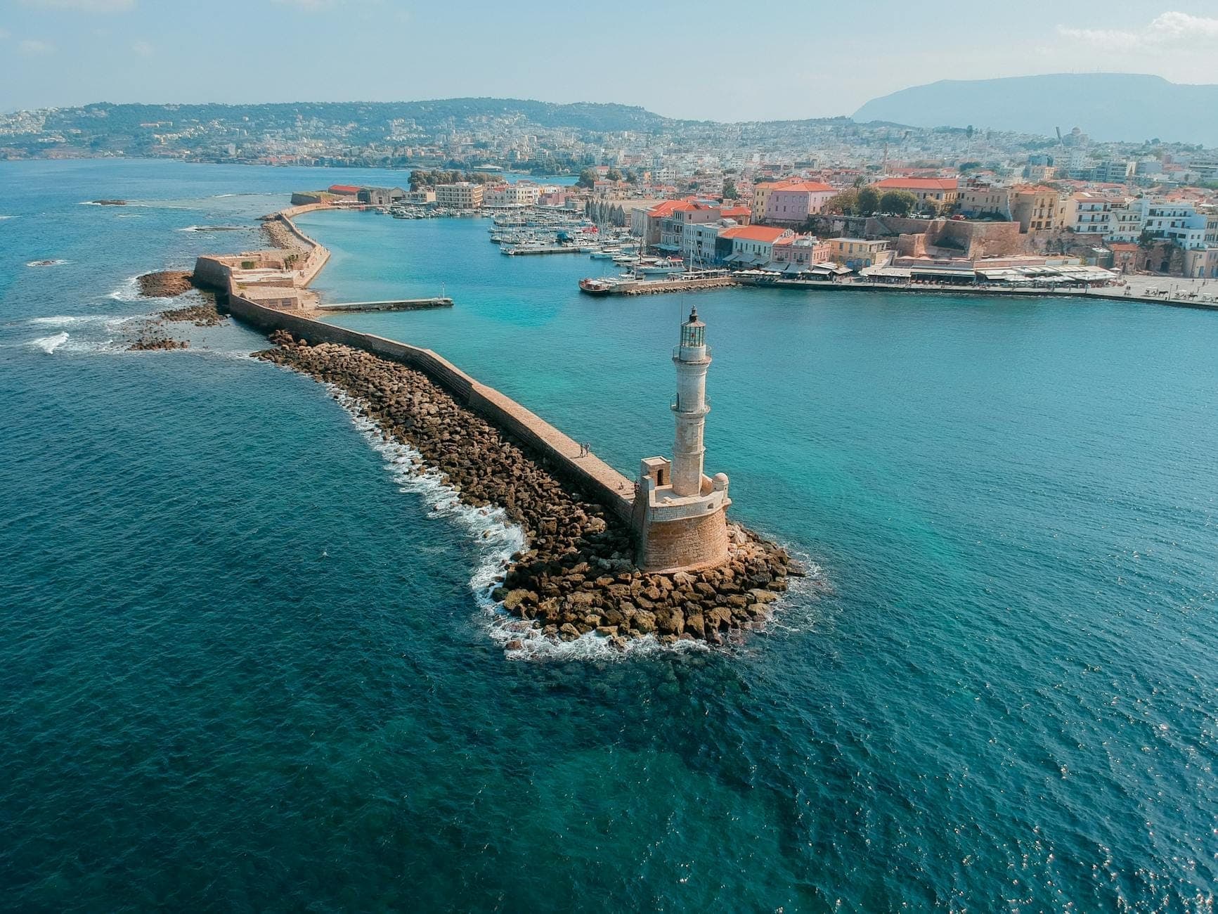 Luftaufnahme des Hafens von Chania auf Kreta mit dem ikonischen Leuchtturm, der langen Seemauer und der farbenfrohen Altstadt vor dem Hintergrund von Bergen.