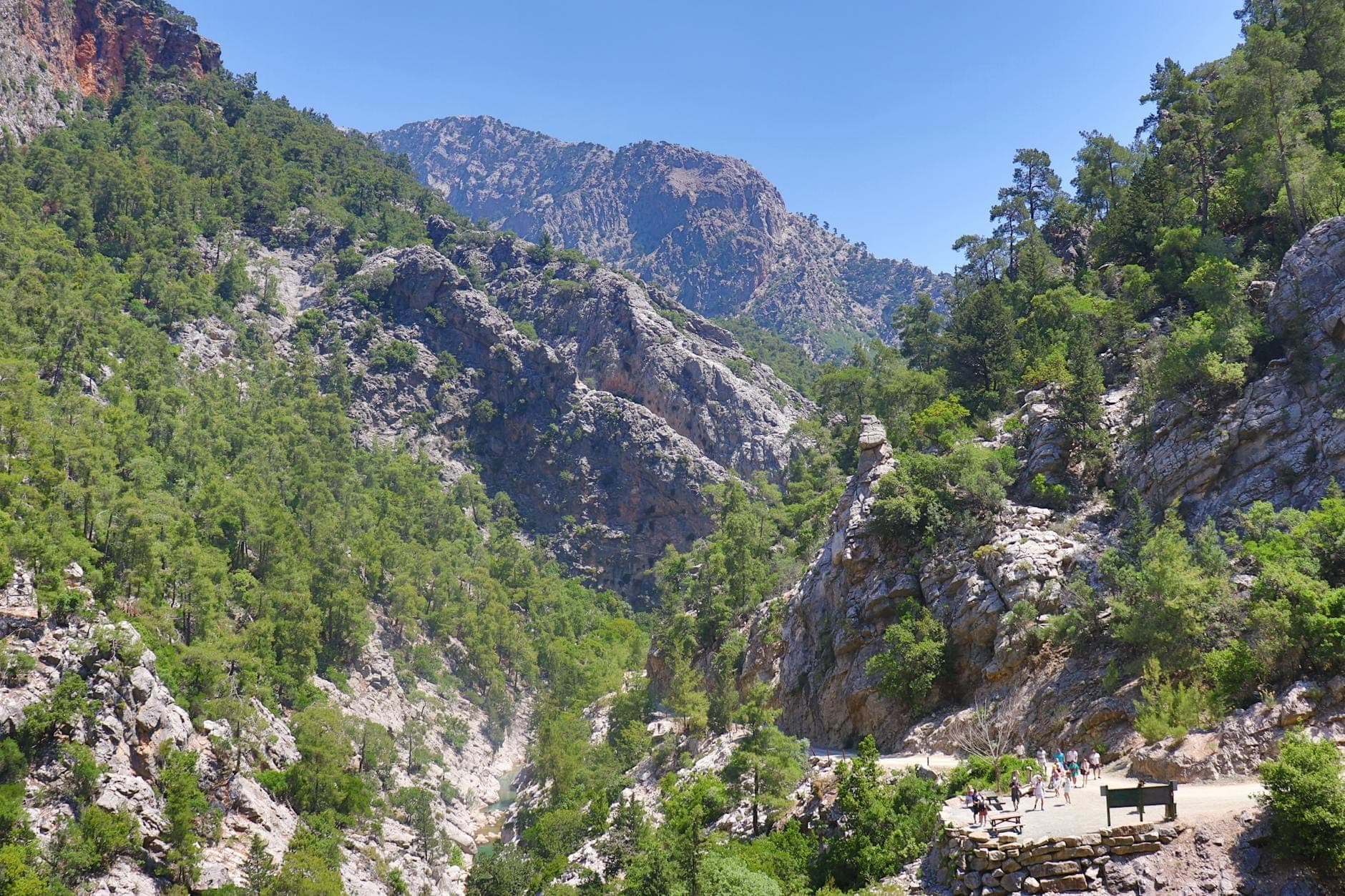Weiter Blick auf eine dramatische Felsschlucht, gesäumt von Kiefern im Lefka Ori auf Kreta, mit Wanderern auf einem malerischen Bergpfad.