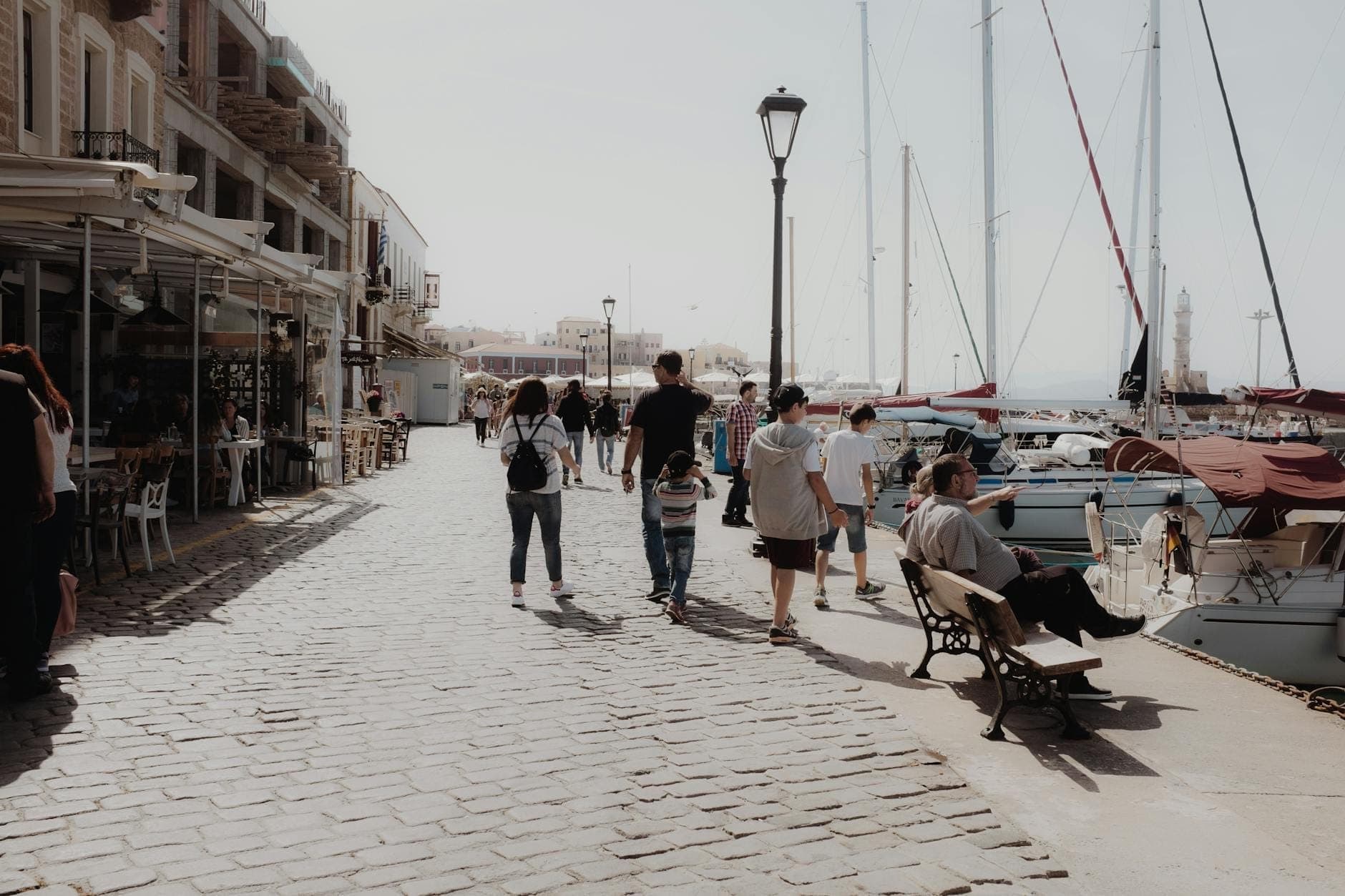Familien und Besucher schlendern entlang einer malerischen Hafenpromenade auf Kreta, mit angedockten Booten und Cafés entlang des Kopfsteinpflasterwegs.