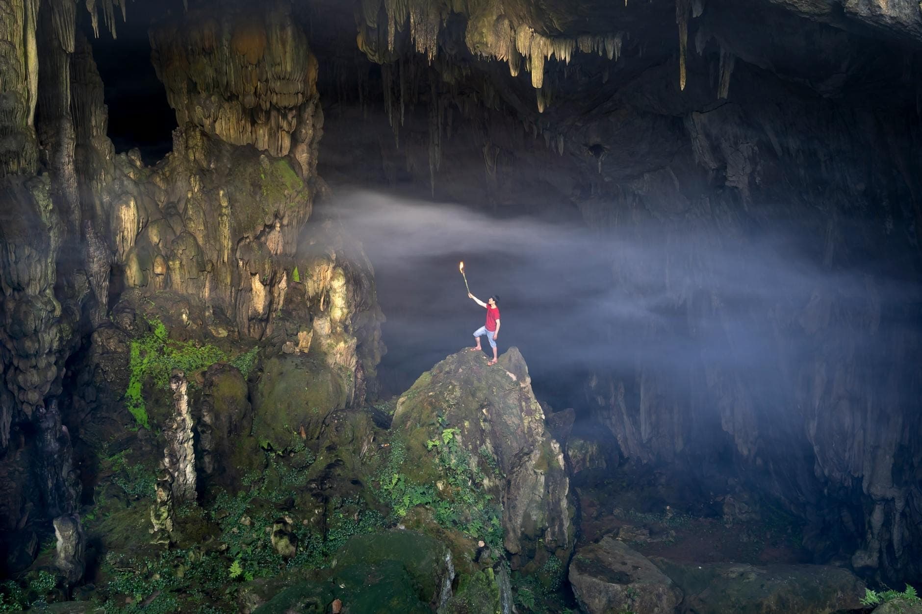 Person mit einer Fackel in einer großen Höhle mit beeindruckenden Stalaktiten, Stalagmiten und einem Sonnenstrahl, der das Innere beleuchtet.