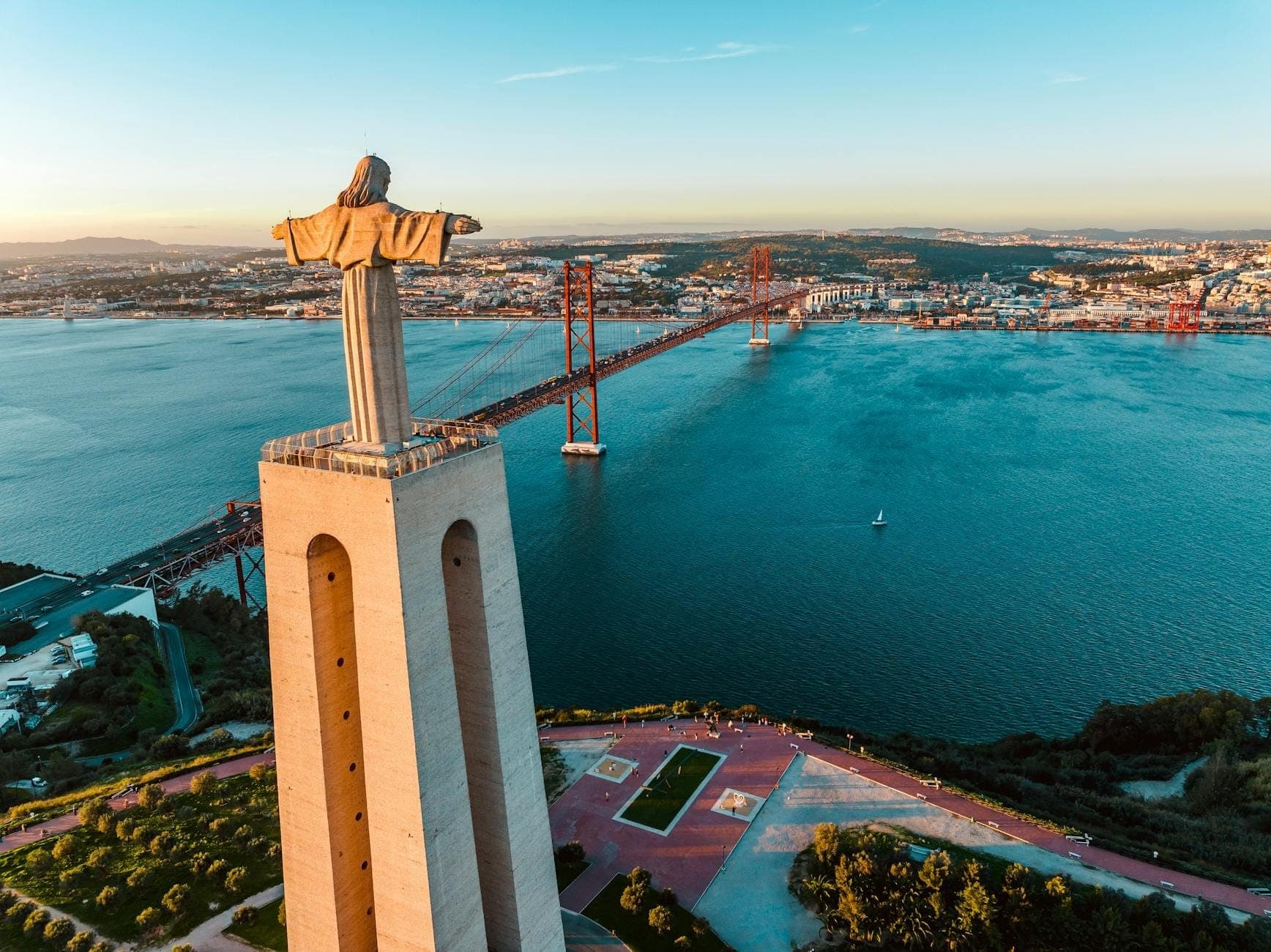 Vista aérea da estátua do Cristo Rei sobre o Rio Tejo e a Ponte 25 de Abril, com Lisboa e a paisagem ao redor sob um céu azul límpido.