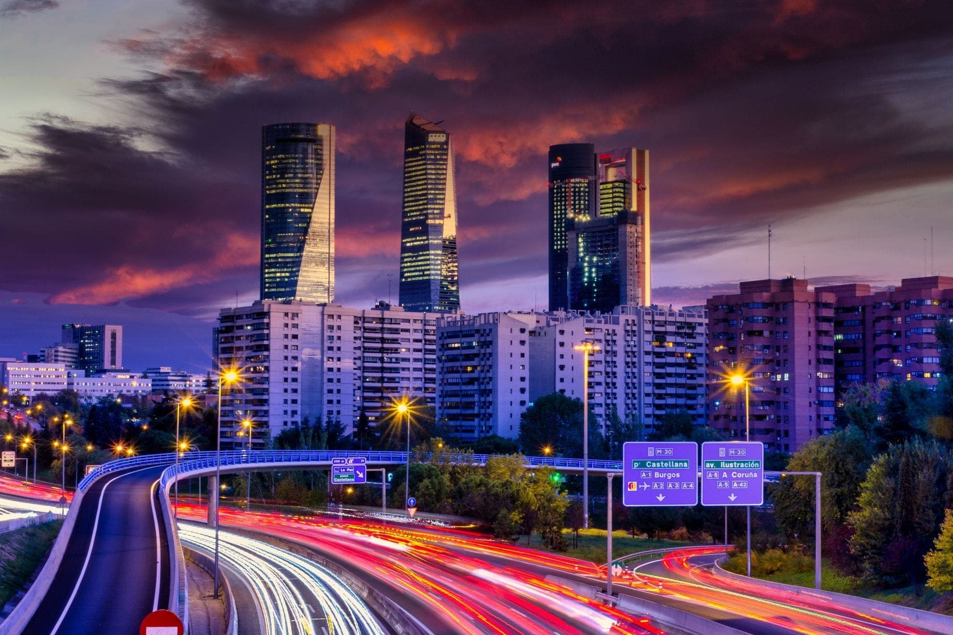 ตึกระฟ้า Cuatro Torres ยามพระอาทิตย์ตก มีเมฆก้อนใหญ่และถนนเมืองมาดริดส่องไฟเป็นทางยาวด้านหน้า