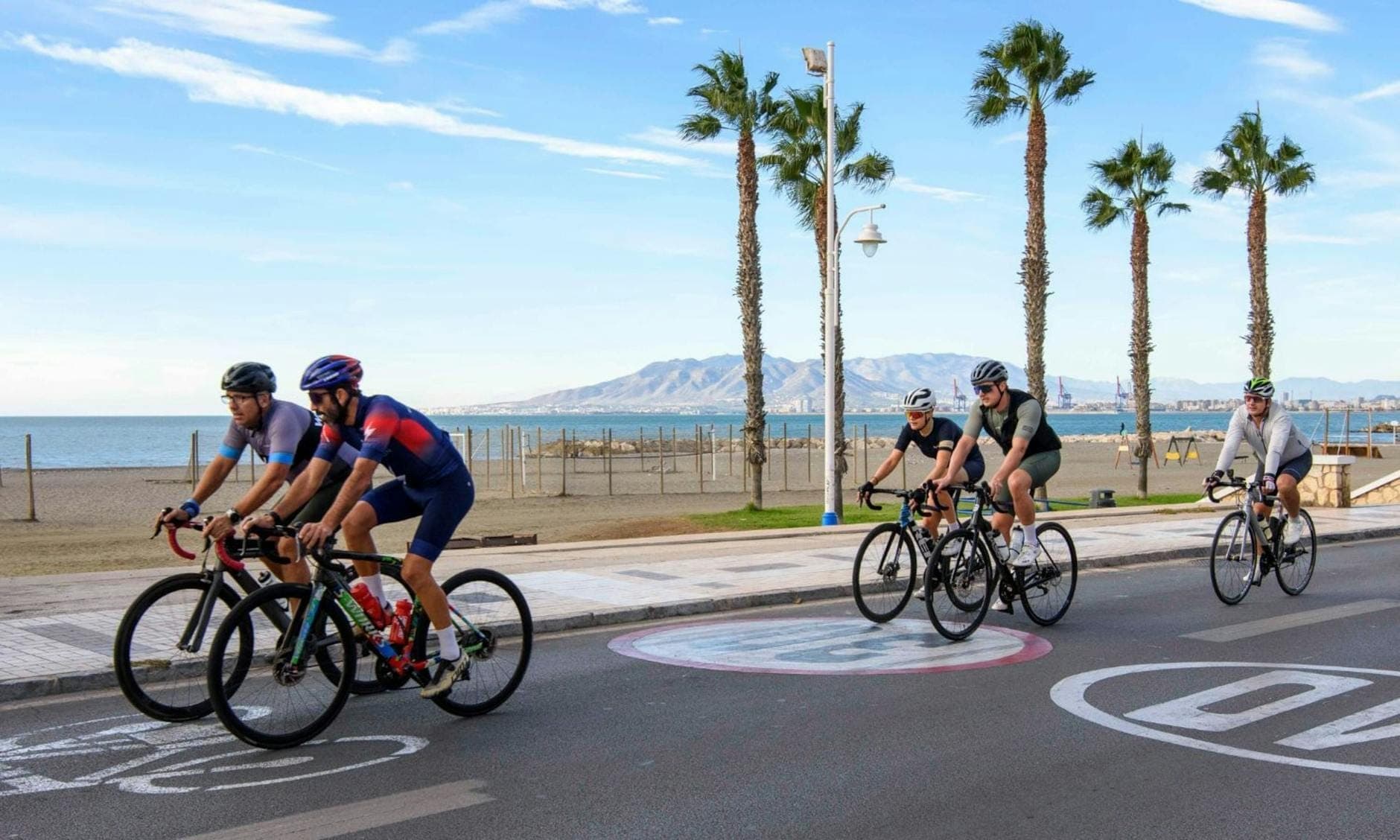 Gruppe von Radfahrern auf einer palmengesäumten Küstenstraße auf Mallorca mit Bergen und blauem Meer im Hintergrund unter klarem Himmel.